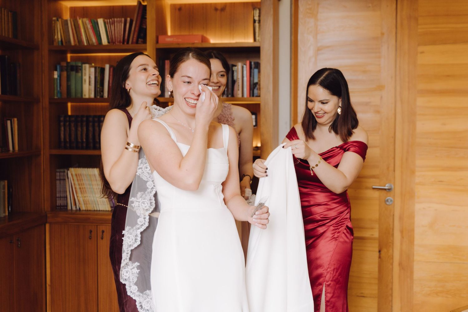 Bridesmaids laughing and assisting emotional bride getting ready in a wood-paneled library room.
