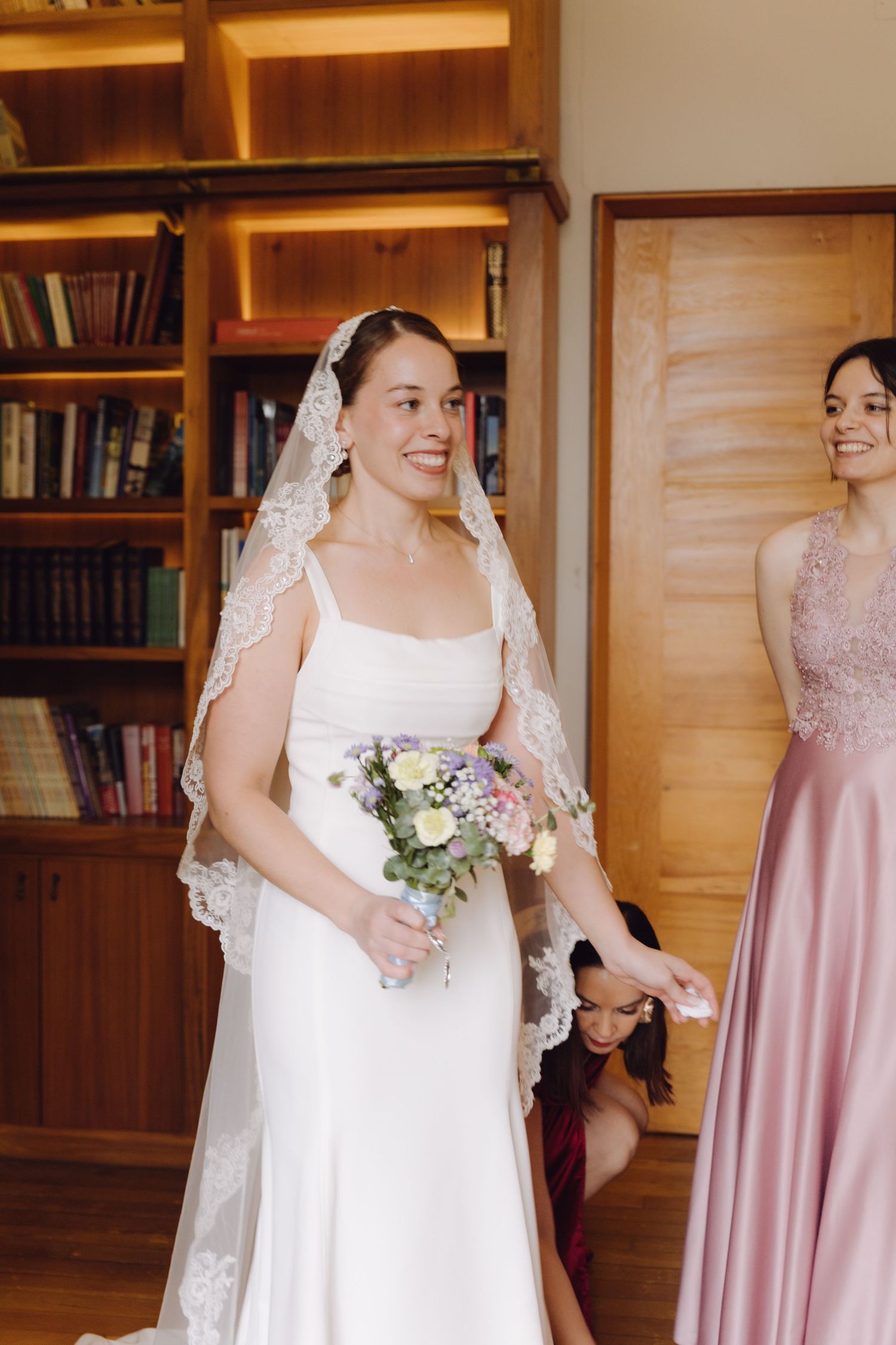 Radiant bride in white gown and lace veil holding pastel bouquet with bridesmaids in library.