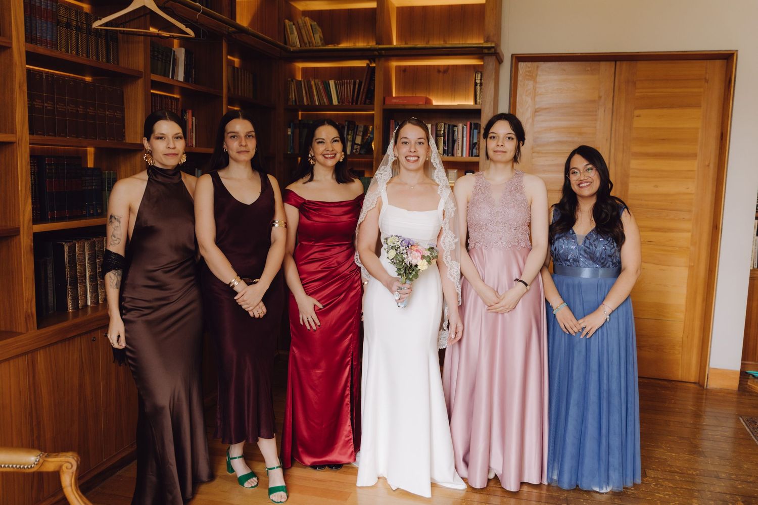Bride in white gown posing with five bridesmaids in colorful dresses in a library setting.