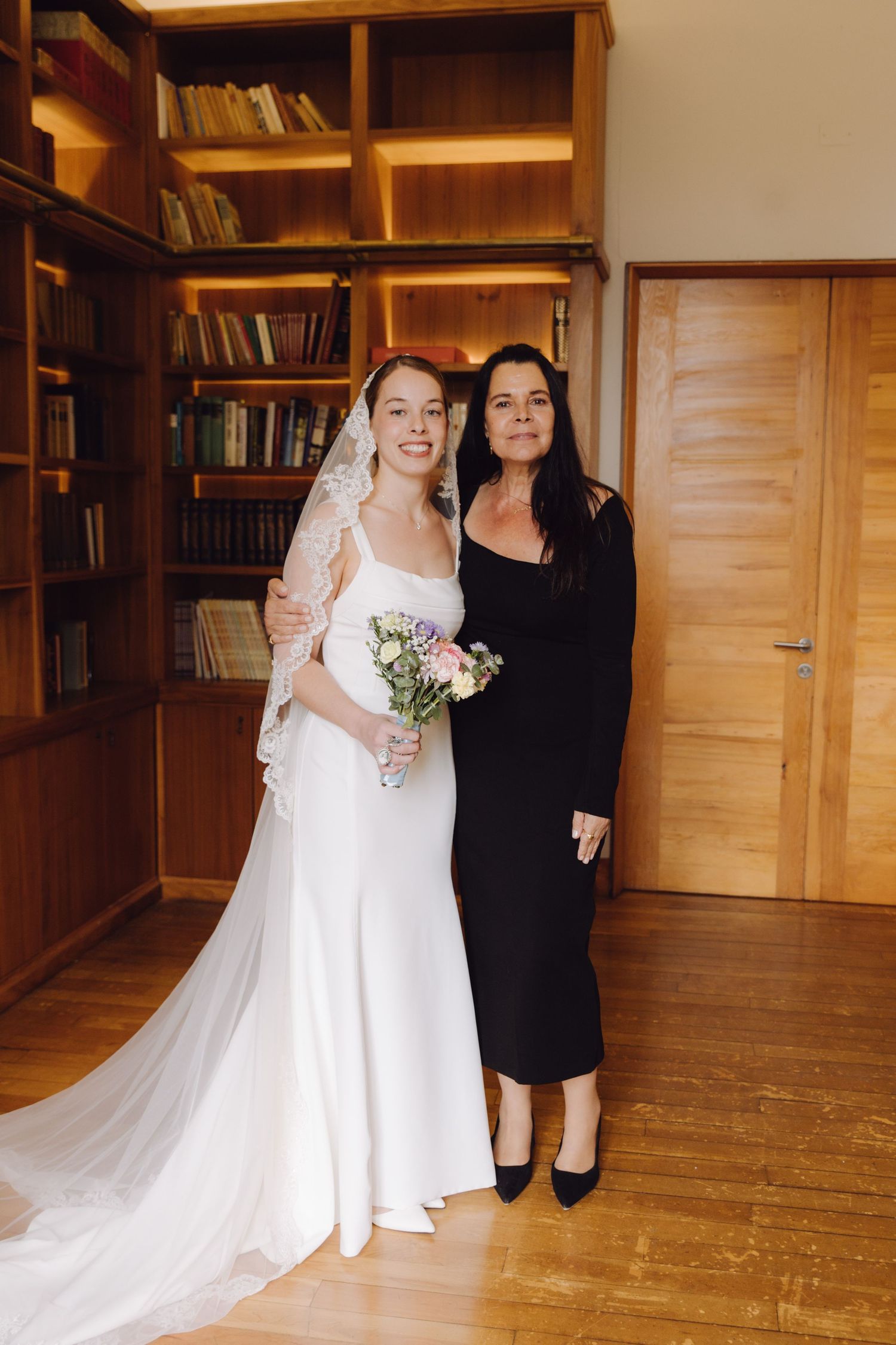 Elegant bride in white gown and lace veil posing with woman in black dress in library.