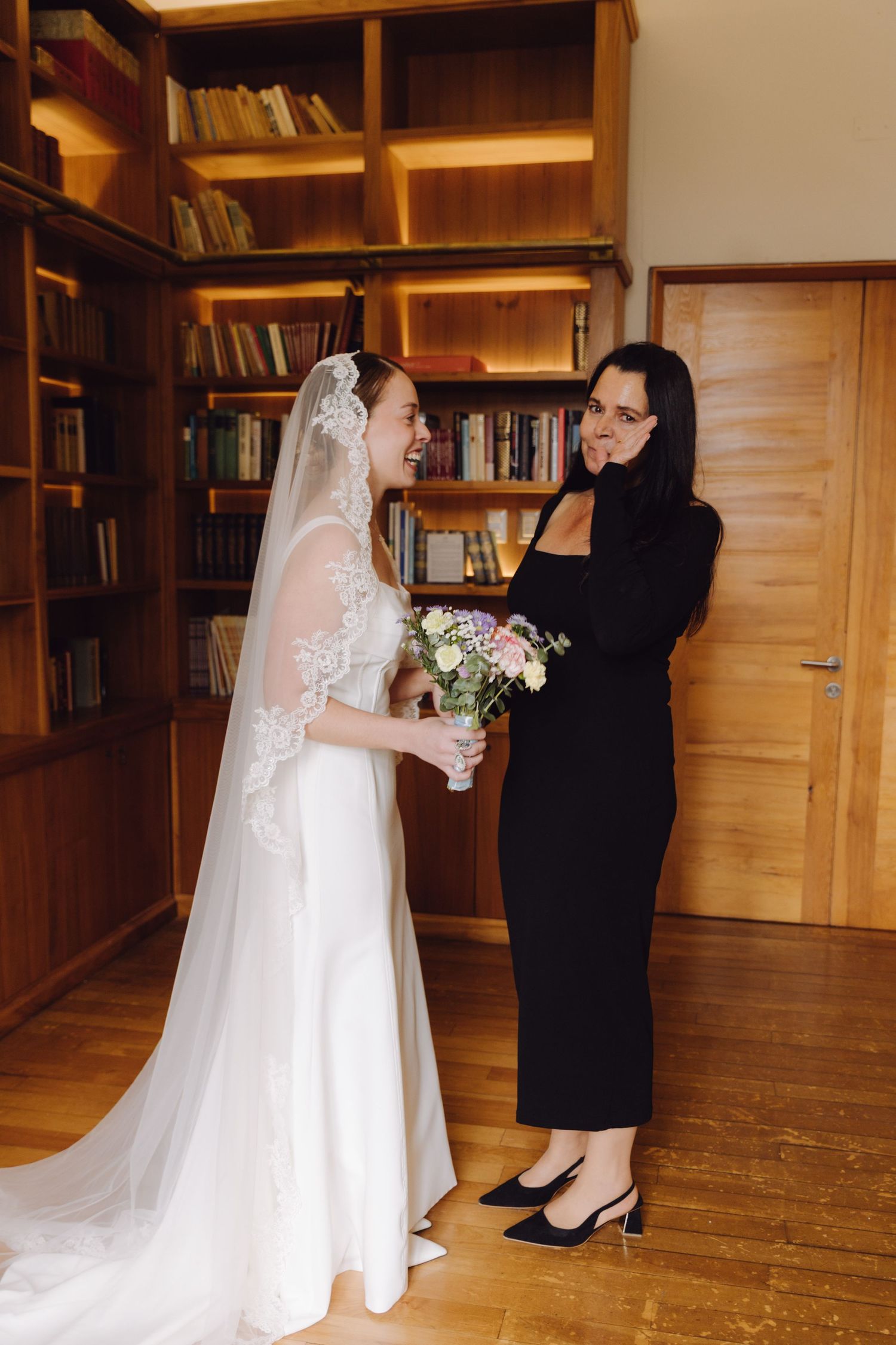 Bride in white lace veil laughing with woman in black dress holding pastel bouquet in library.