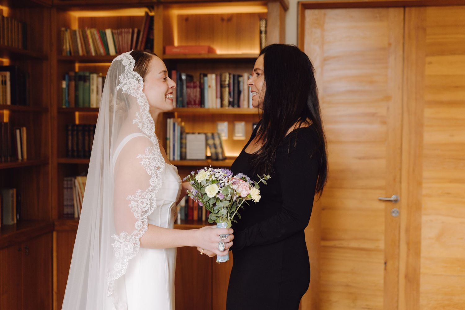 Bride in white lace veil smiling at woman in black dress while holding pastel bouquet indoors.
