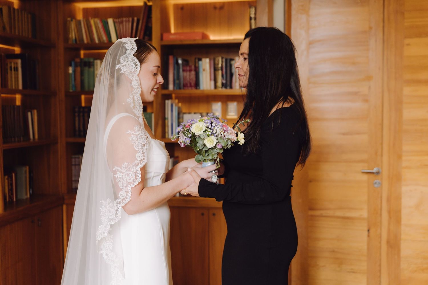 Bride in white gown and lace veil holds bouquet while woman in black dress holds her hands in a library.
