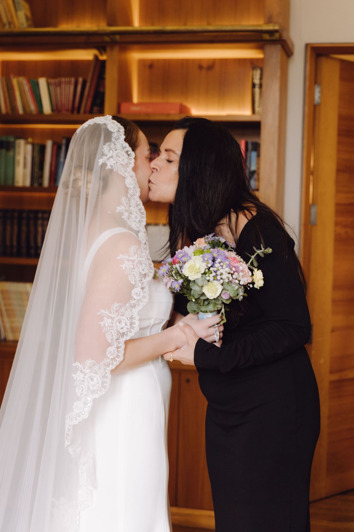 Bride in white wedding gown and lace veil shares a kiss with woman in black dress holding a floral bouquet.
