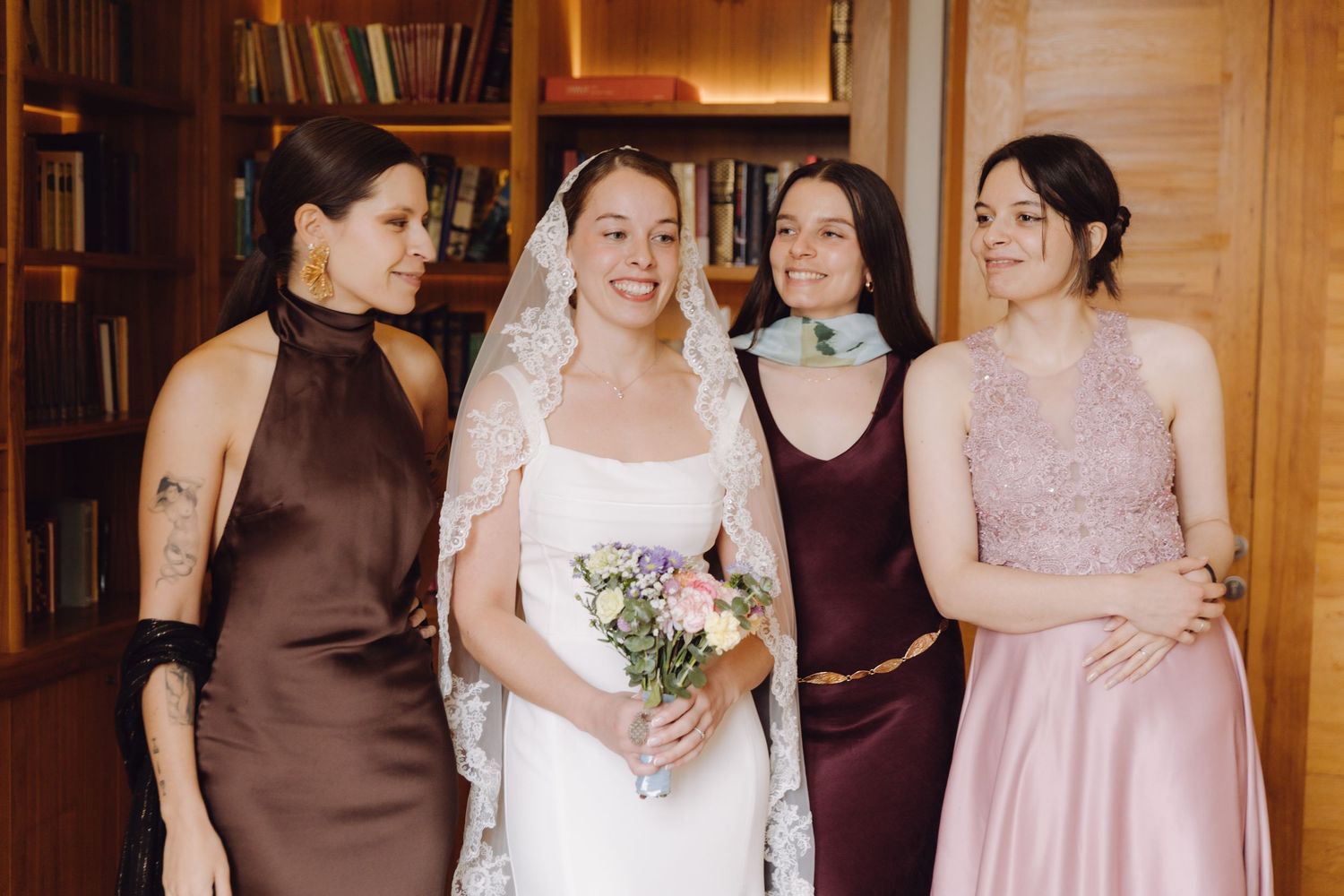 Smiling bride in white dress and lace veil poses with three bridesmaids in colorful gowns in a library.