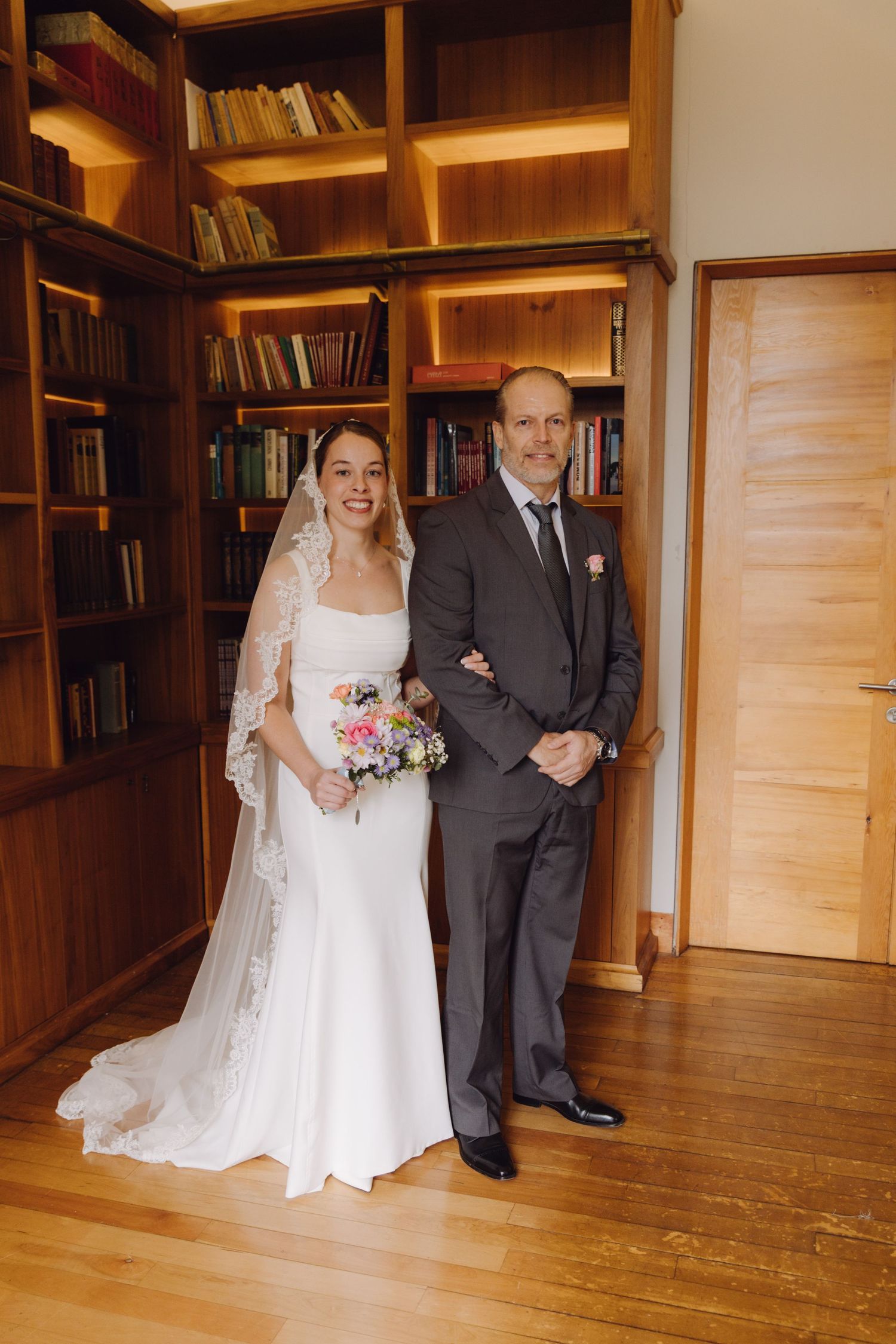 Bride in white gown and lace veil stands with older man in gray suit in front of wooden bookshelves.