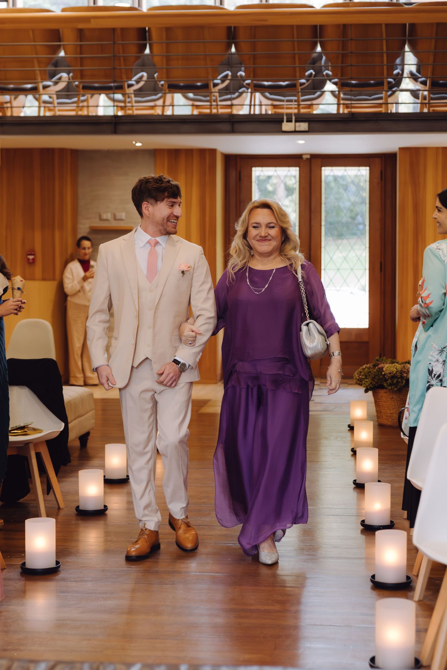 Groom in beige suit with pink tie walks down candlelit aisle with woman in purple dress at wedding.
