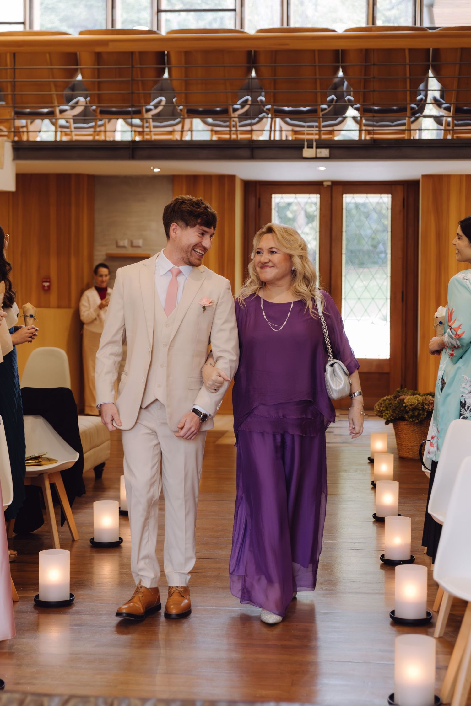 Smiling groom in cream suit escorts woman in purple gown down a candlelit wedding ceremony aisle.