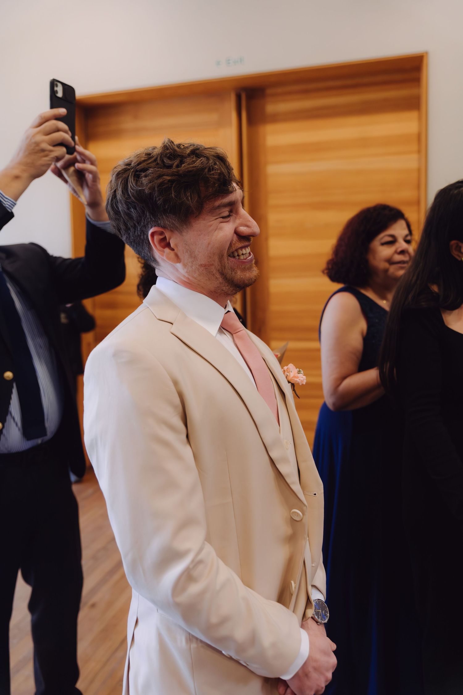 Emotional groom in beige suit laughs joyfully as he watches bride walk down the wedding aisle.