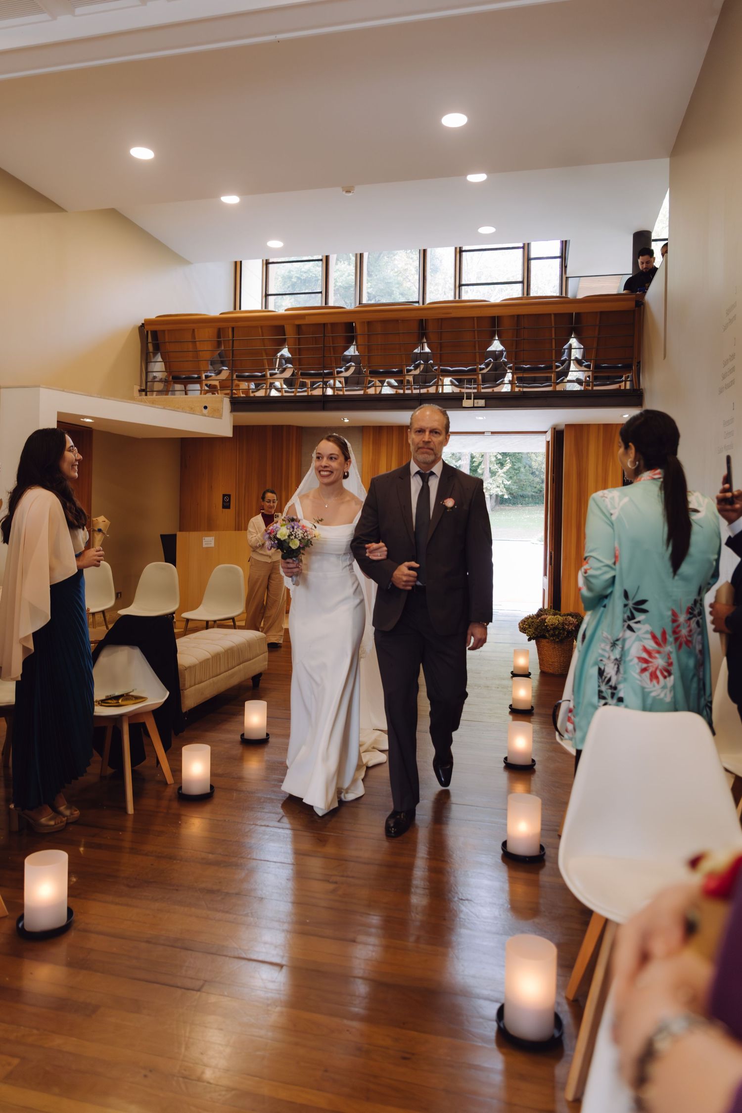 Bride in white gown and veil walks down candlelit aisle escorted by man in dark suit at ceremony.