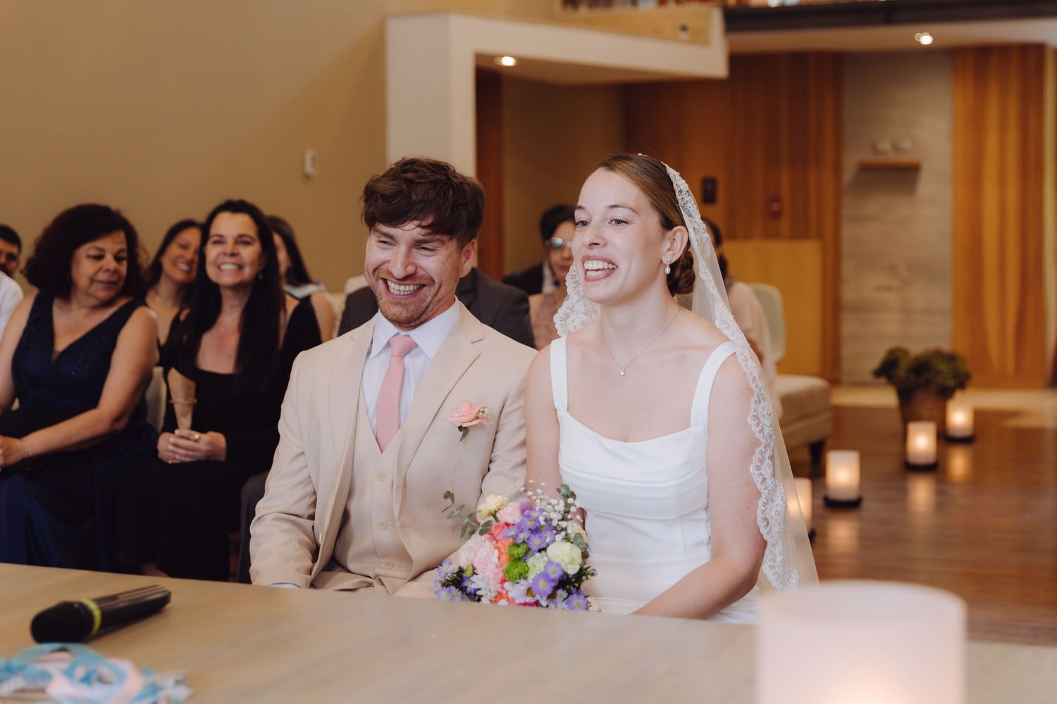 Bride and groom sit together laughing at the altar during their wedding ceremony with guests behind.
