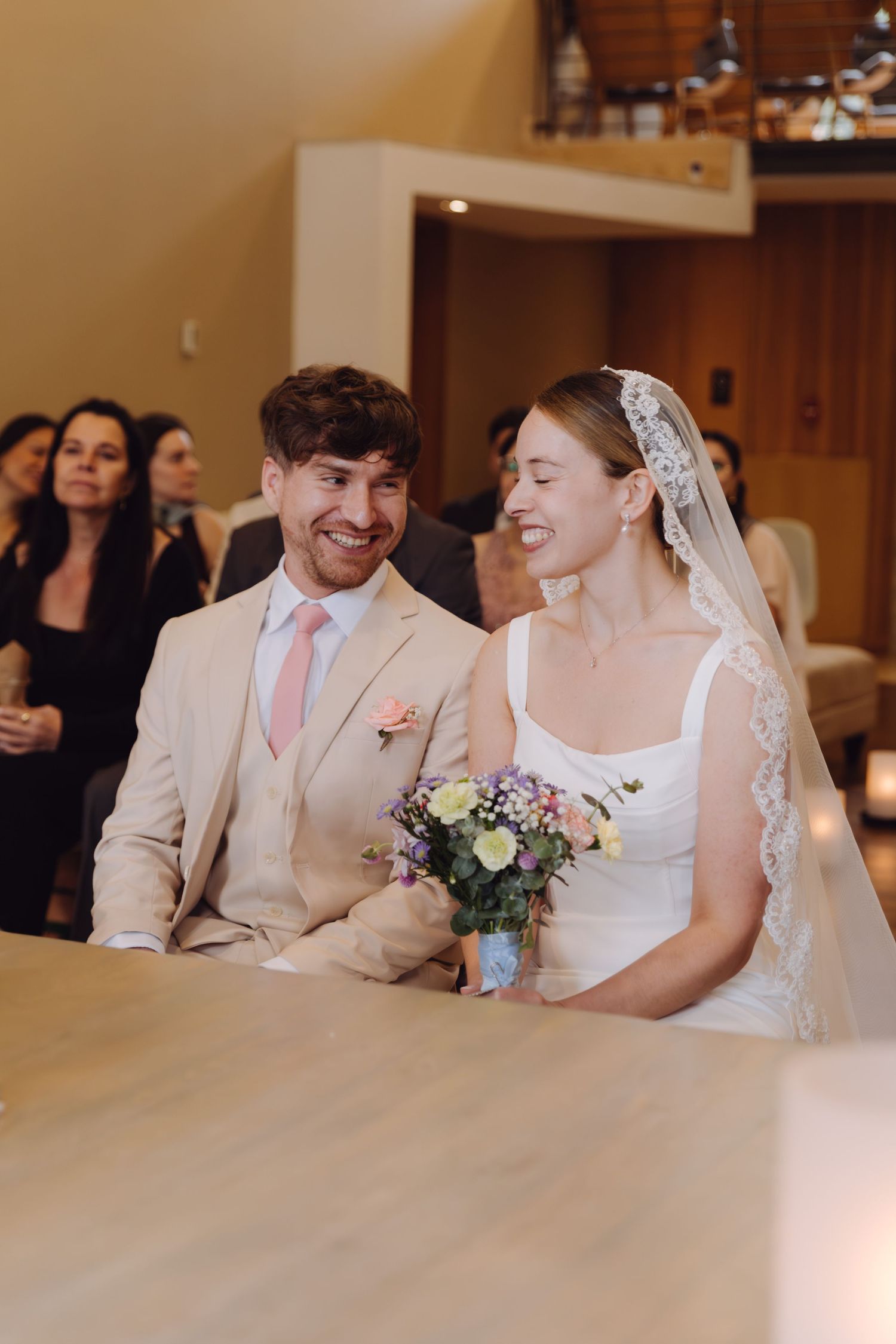 Smiling bride and groom sitting together at wedding ceremony altar, bride holding colorful bouquet.