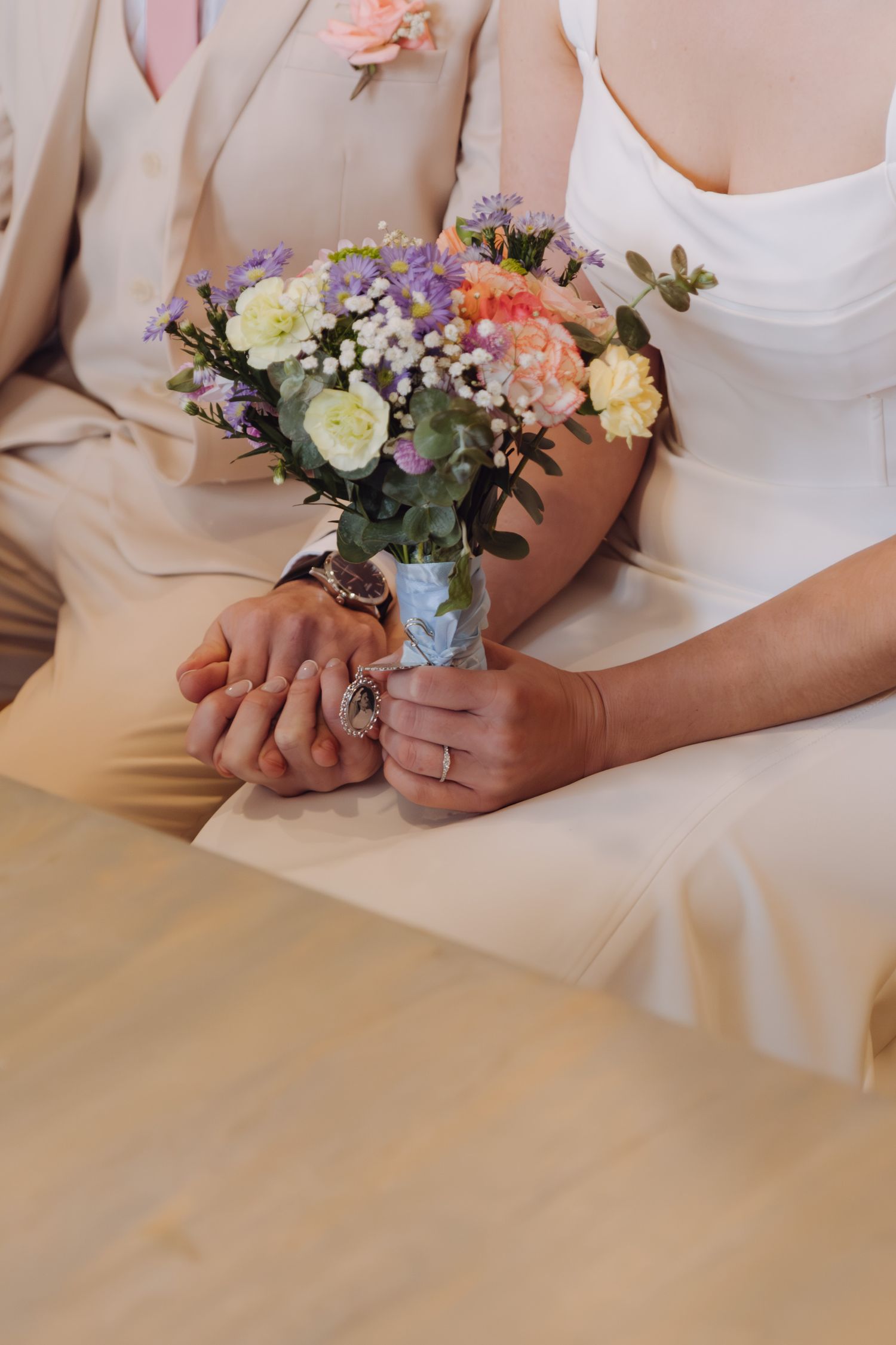 Close-up of bride and groom holding hands with colorful wedding bouquet during ceremony.
