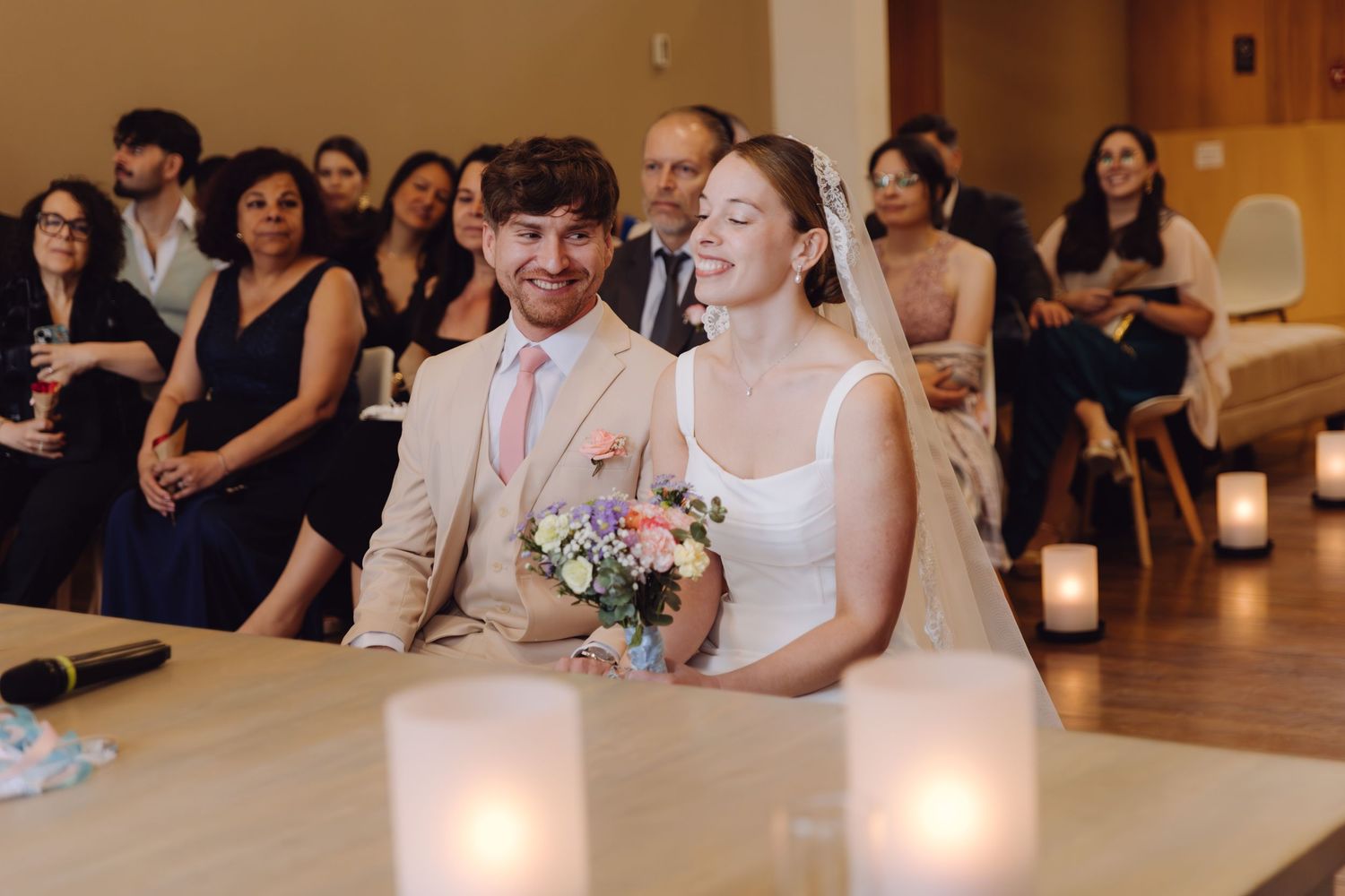 Happy couple laughing together at wedding ceremony table with candles and guests in background.