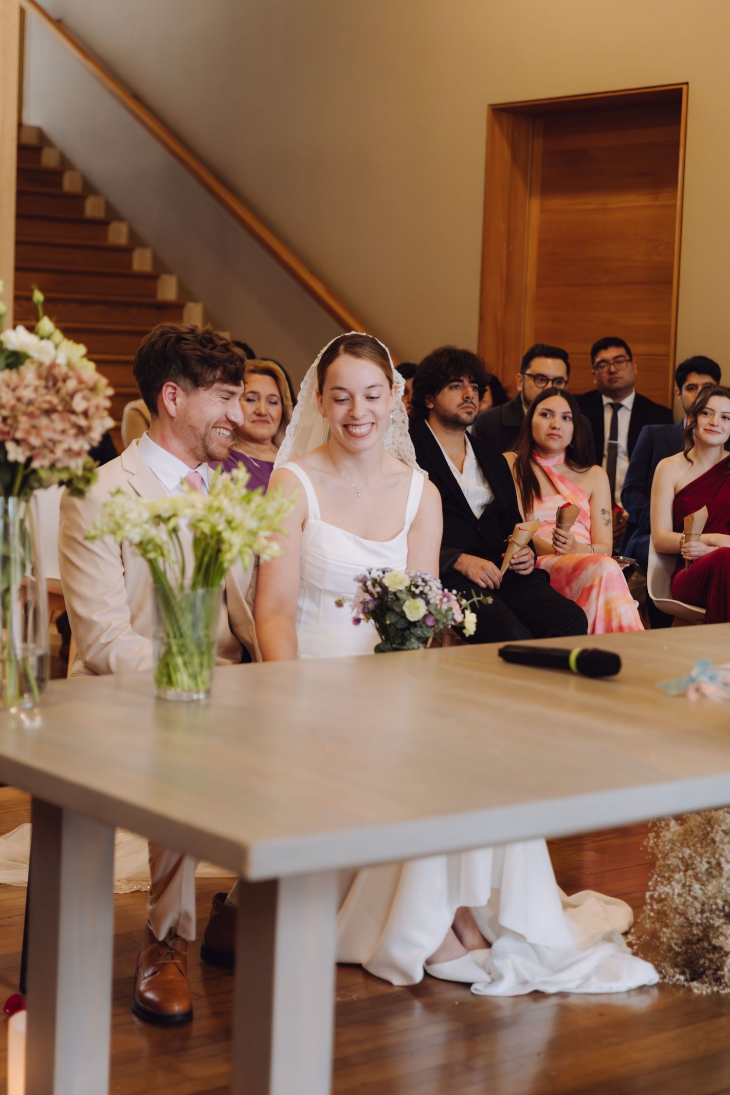Bride and groom smiling at wedding ceremony table with floral arrangements and guests seated behind.