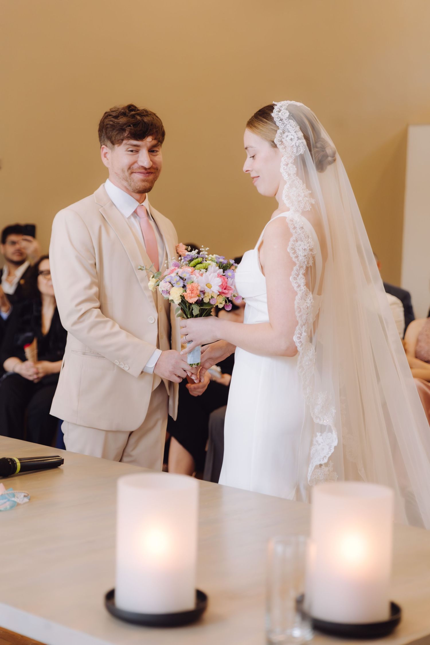 Groom placing wedding ring on bride's finger during indoor ceremony with candles in foreground.