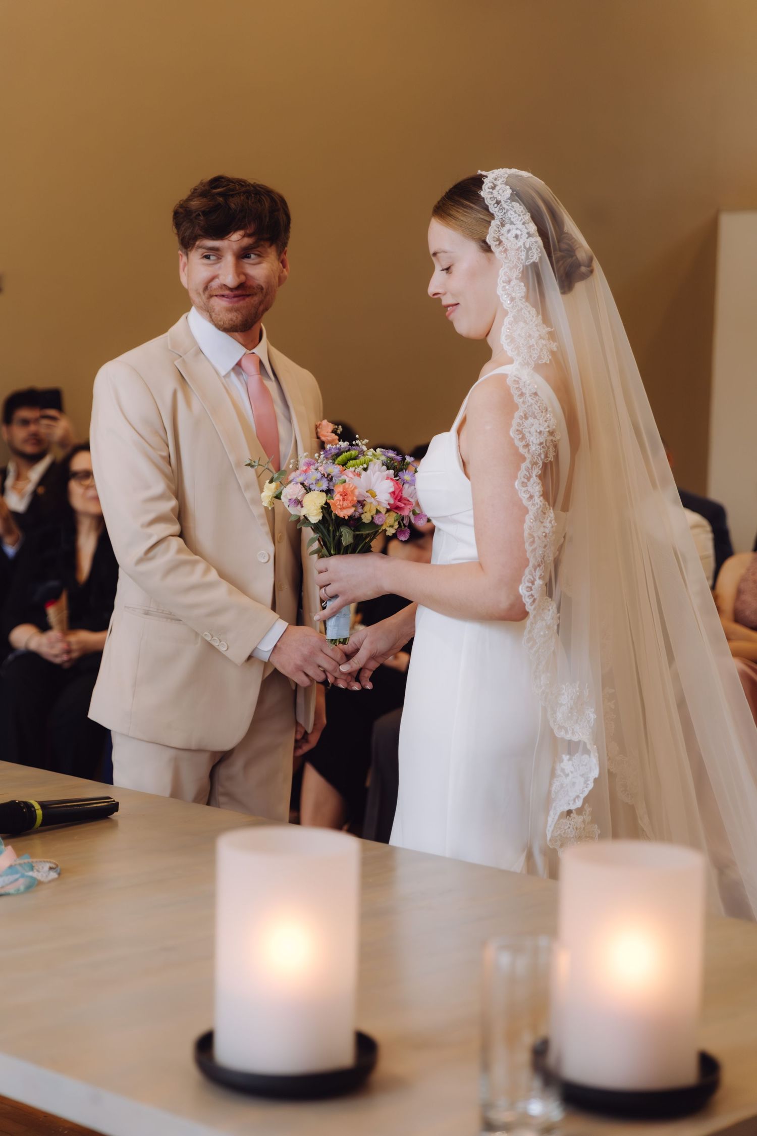 Bride and groom exchanging rings at wedding ceremony with lit candles and guests in background.