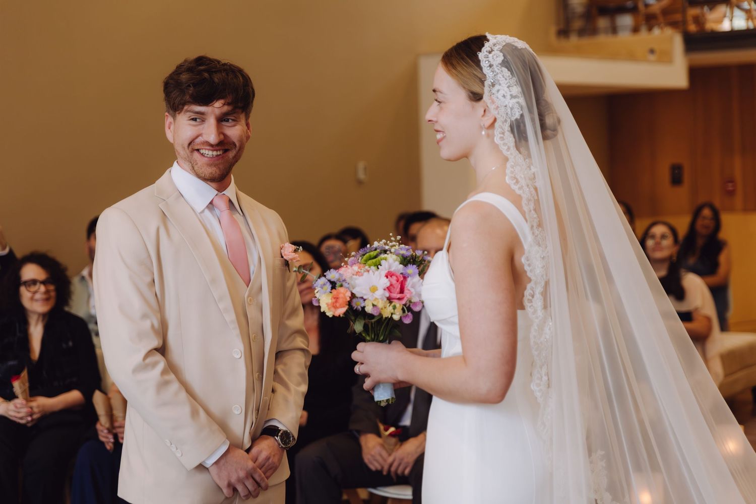 Groom smiling at bride during wedding ceremony, bride holding colorful bouquet with guests behind.