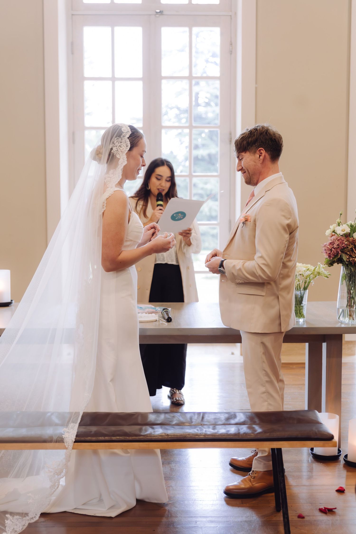 Bride reading vows to groom during wedding ceremony with officiant and large windows behind.