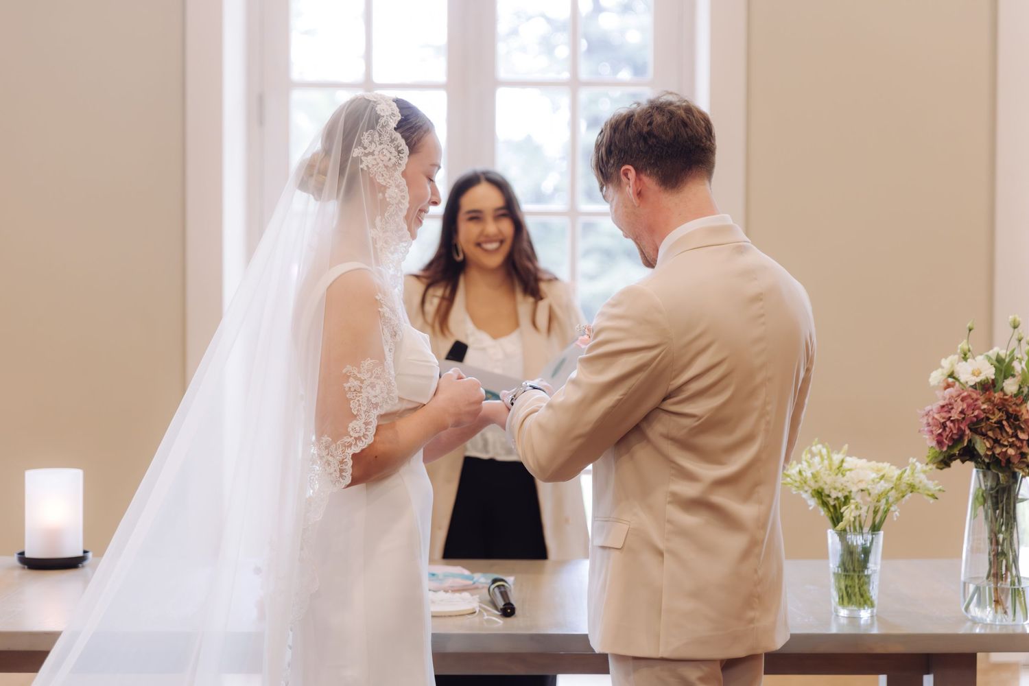Bride and groom exchanging rings with smiling officiant and floral arrangements in background.