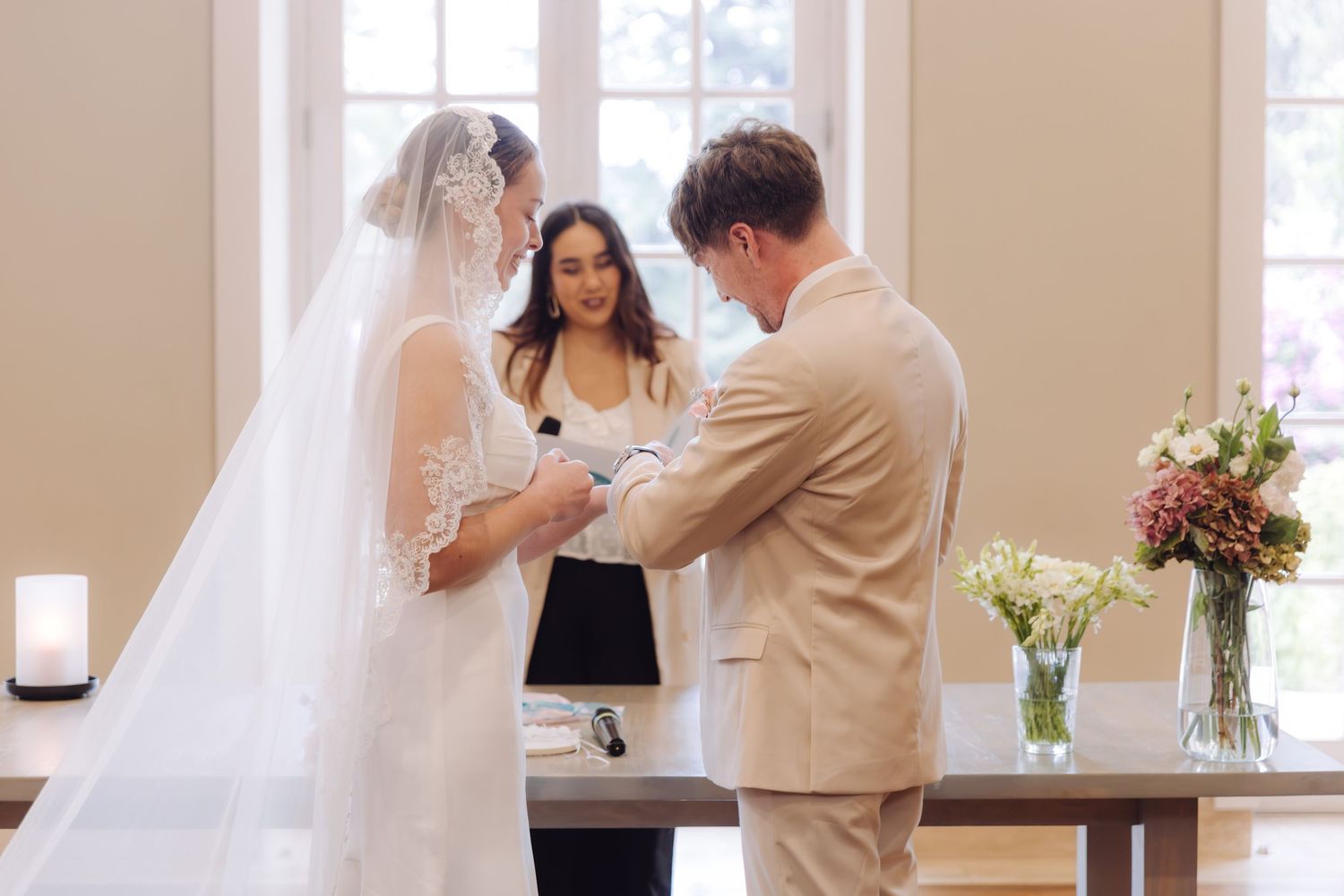 Bride and groom exchange rings during intimate wedding ceremony with smiling officiant.