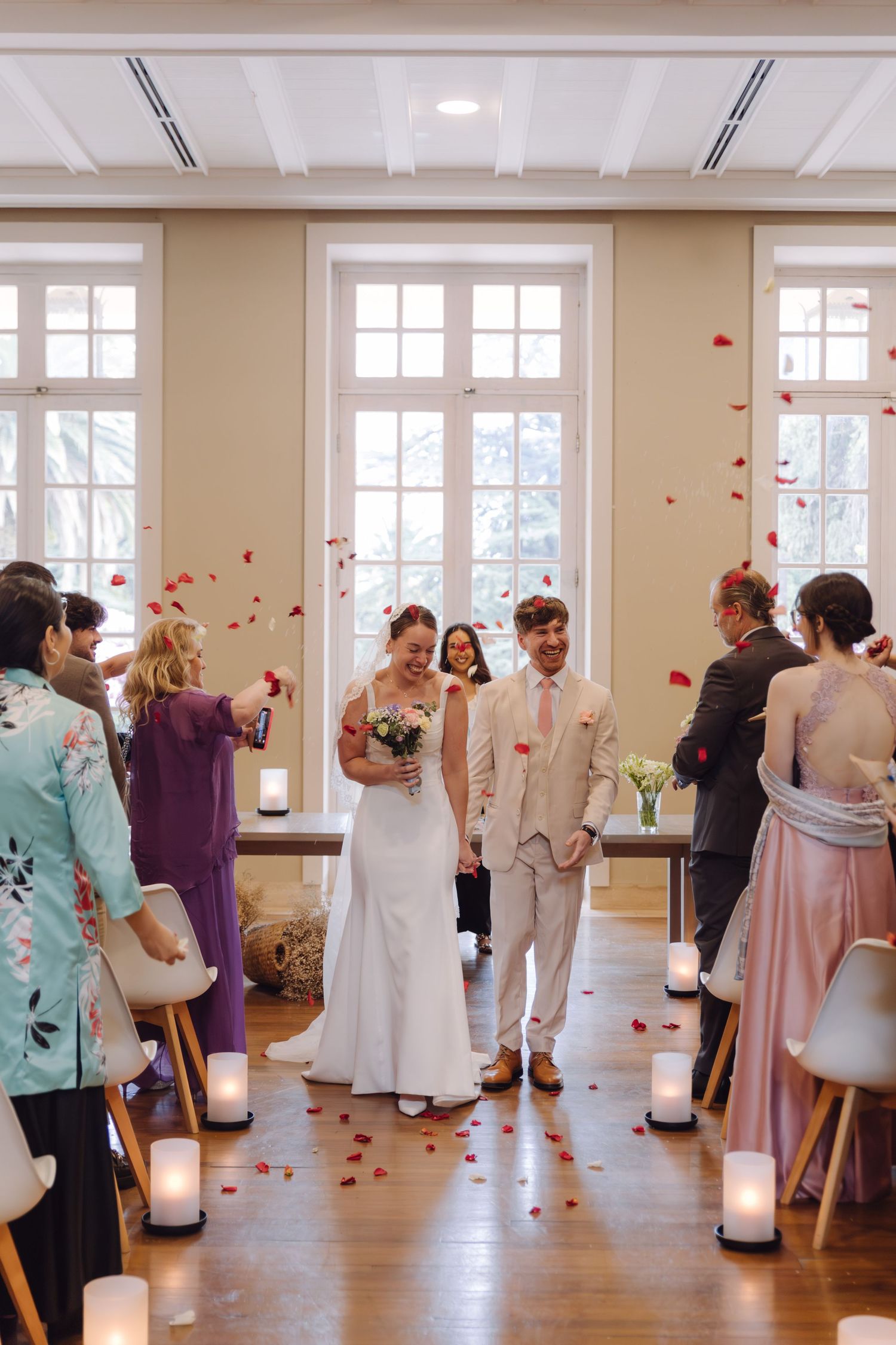 Newlyweds walk down the aisle as guests toss red rose petals in elegant indoor wedding ceremony.