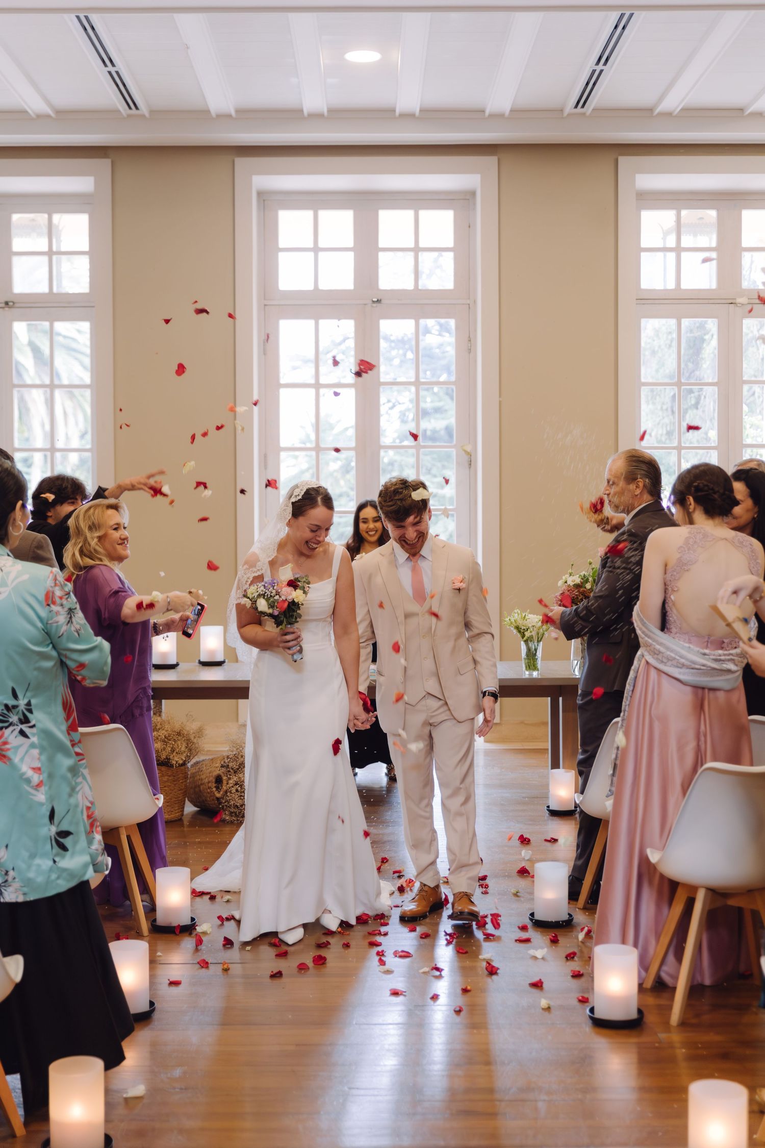 Bride and groom recessional with rose petal confetti thrown by smiling wedding guests indoors.