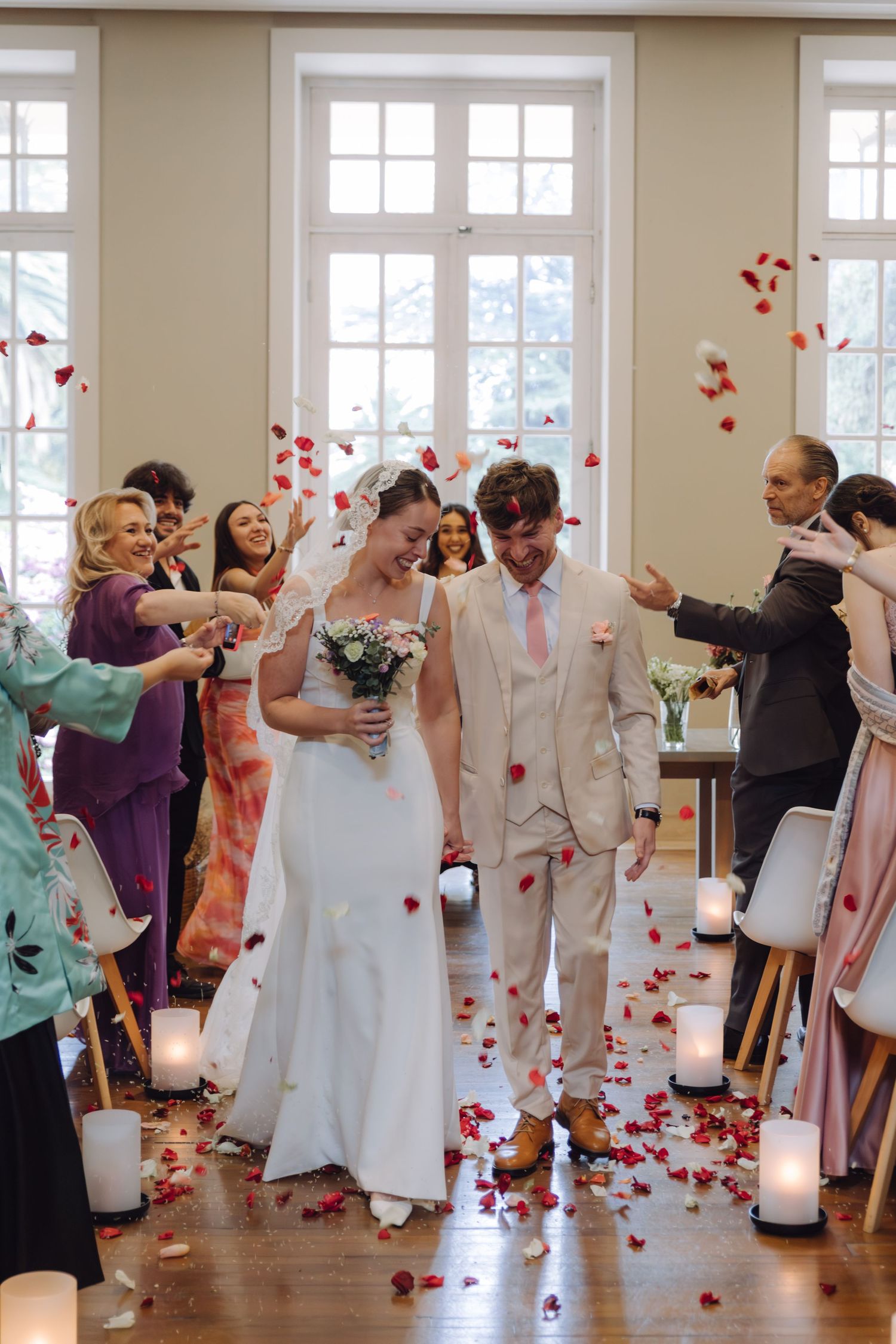 Laughing newlyweds showered with red rose petals by guests during joyful wedding ceremony exit.