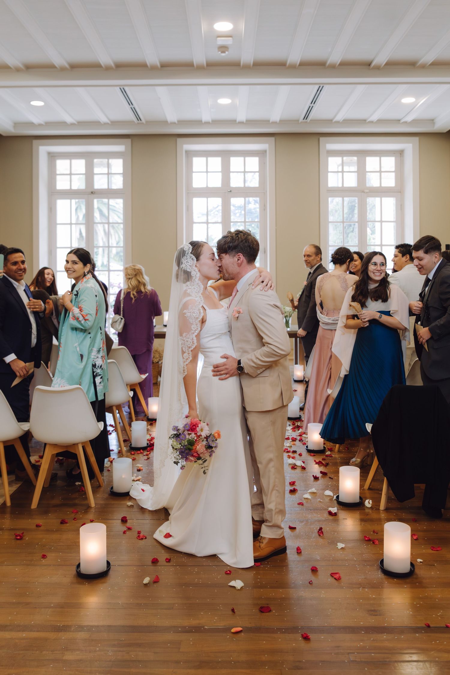 Newlyweds share romantic kiss in aisle after indoor wedding ceremony with rose petals on floor.