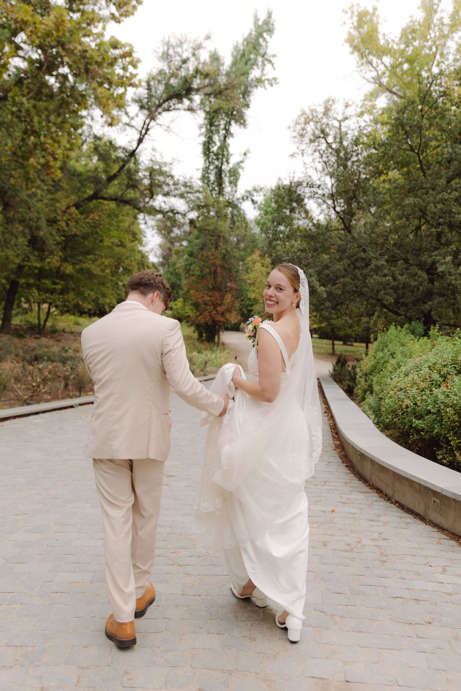 Smiling bride in white gown holds groom's hand while walking on garden path after wedding.