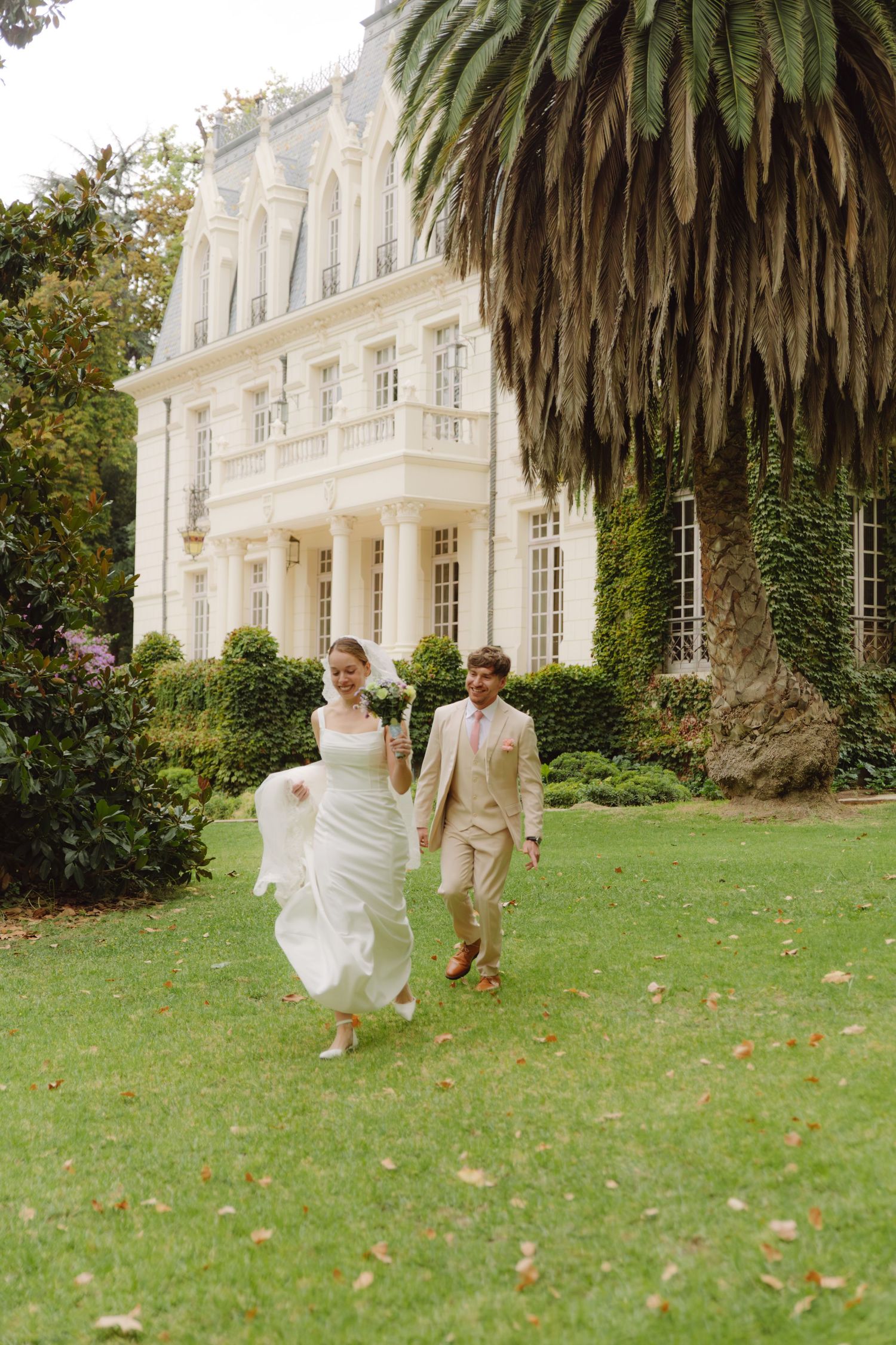 Bride and groom walk across lush lawn in front of elegant white mansion on wedding day.