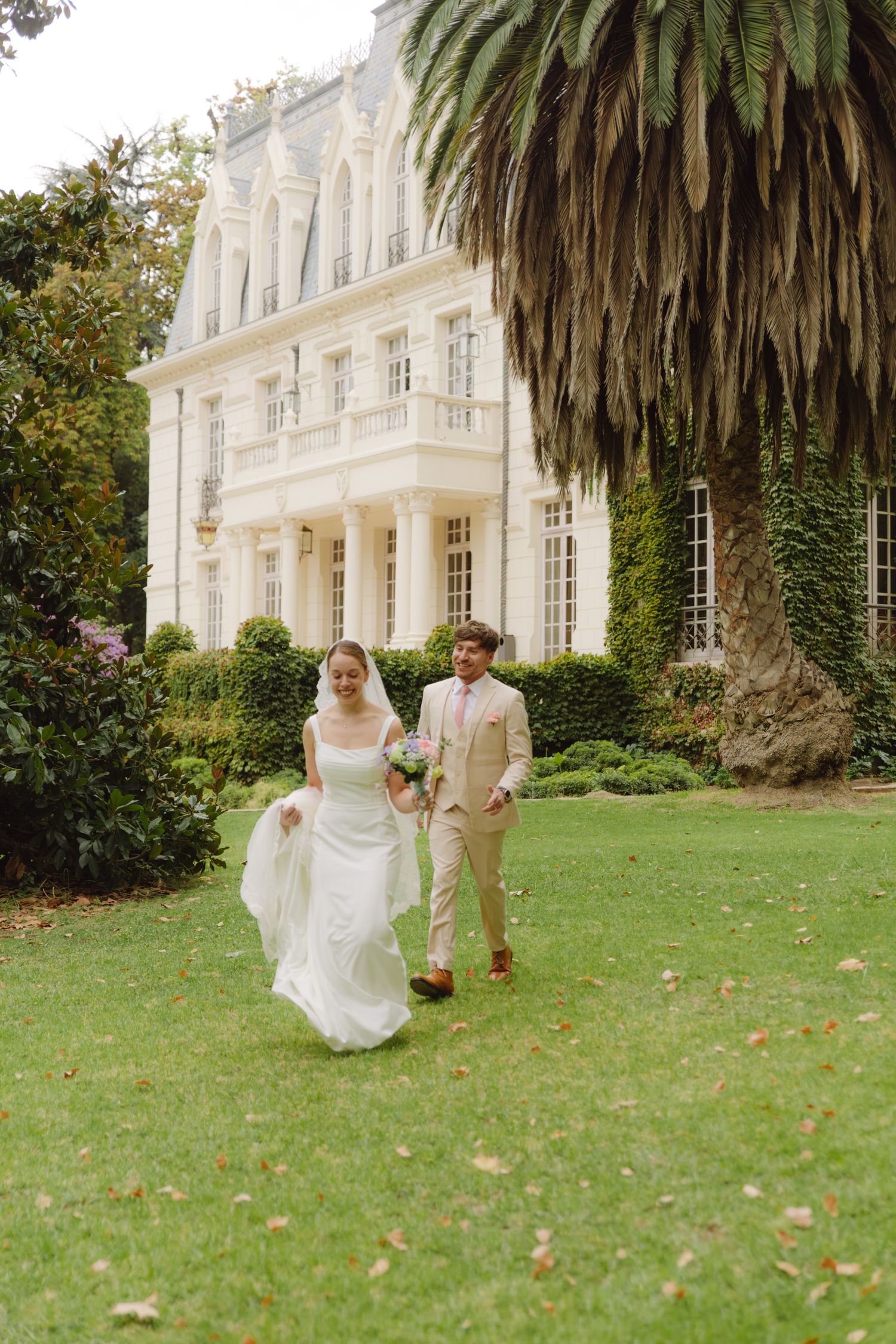 Newlyweds stroll across green lawn with grand white chateau and tall palm tree backdrop.