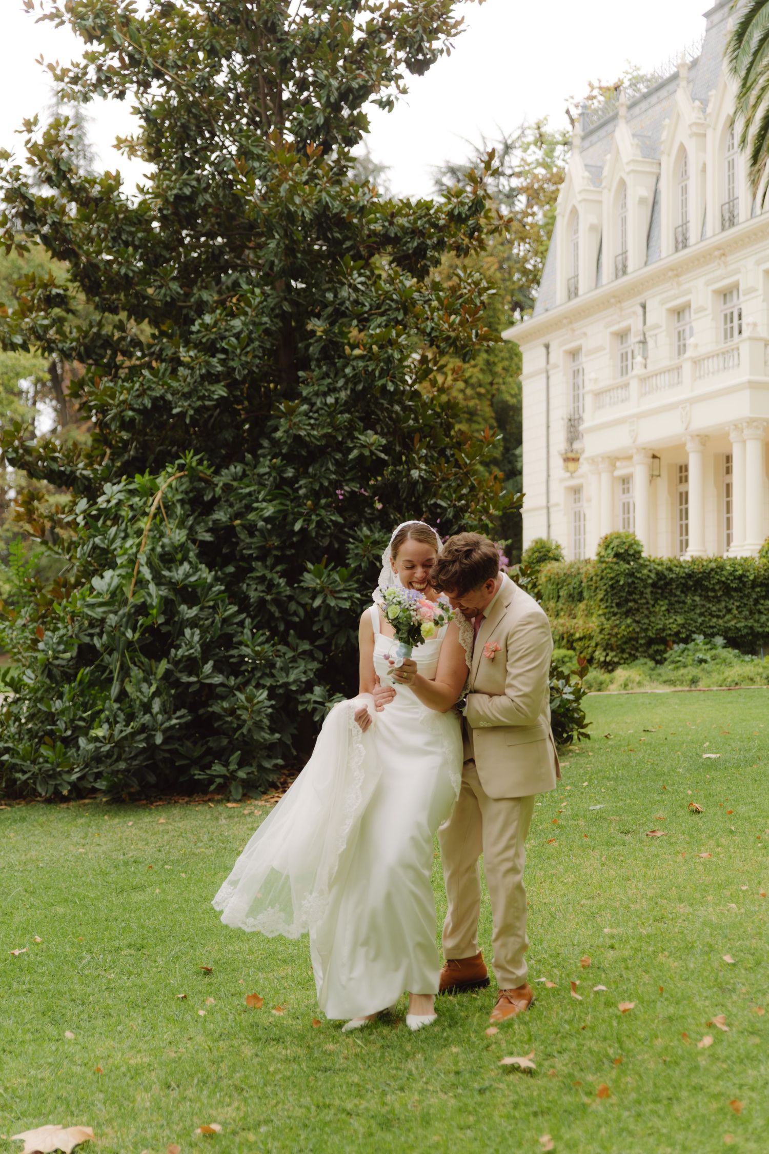 Groom embraces laughing bride holding bouquet on green lawn beside elegant white wedding venue.