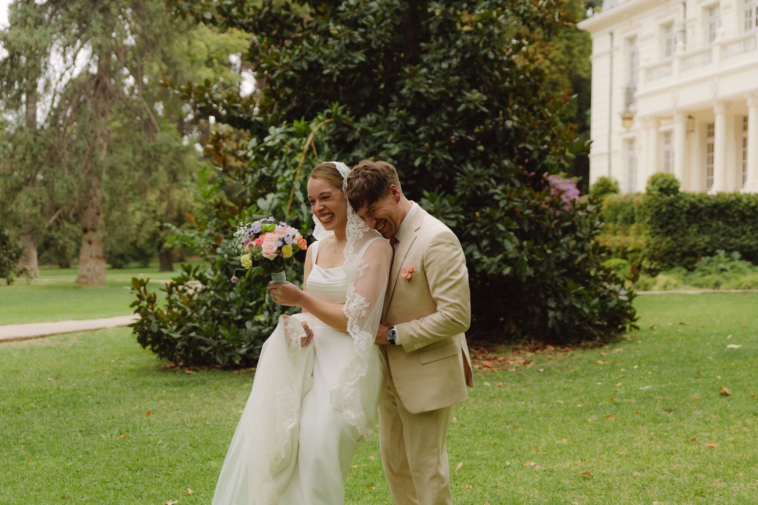 Bride and groom share a joyful moment on green lawn outside elegant white manor house.