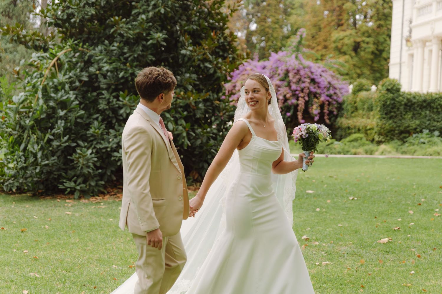 Smiling bride in white gown holds bouquet while walking with groom in beige suit outdoors.