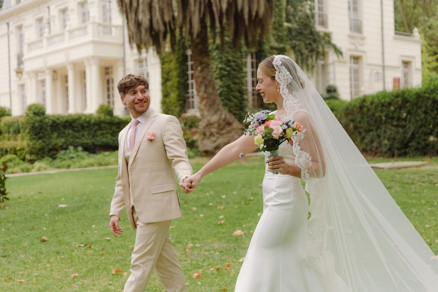 Newlyweds hold hands and walk through a sunlit garden near a grand white estate.