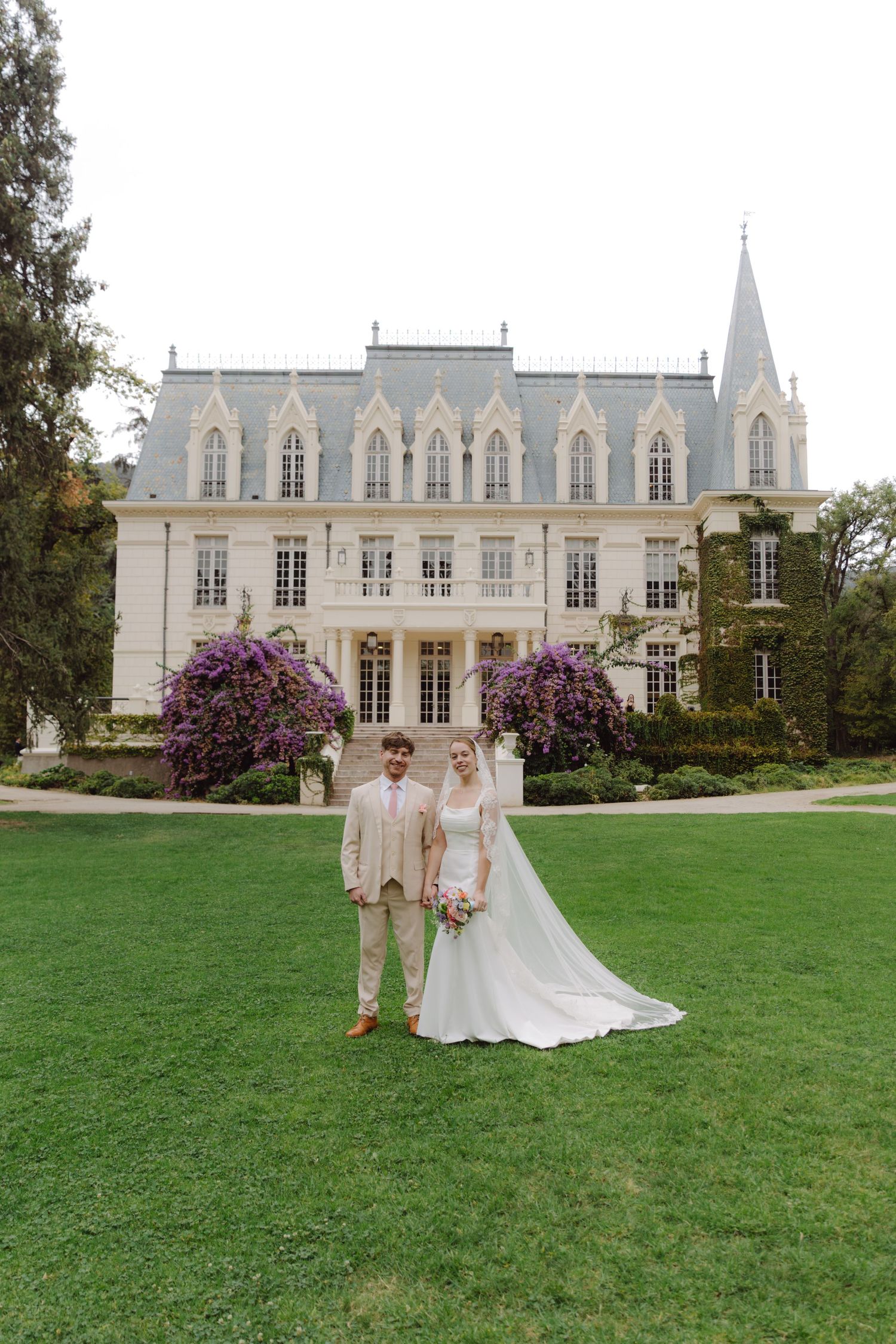 Bride and groom pose before a stunning Gothic-style chateau with purple flowering trees.