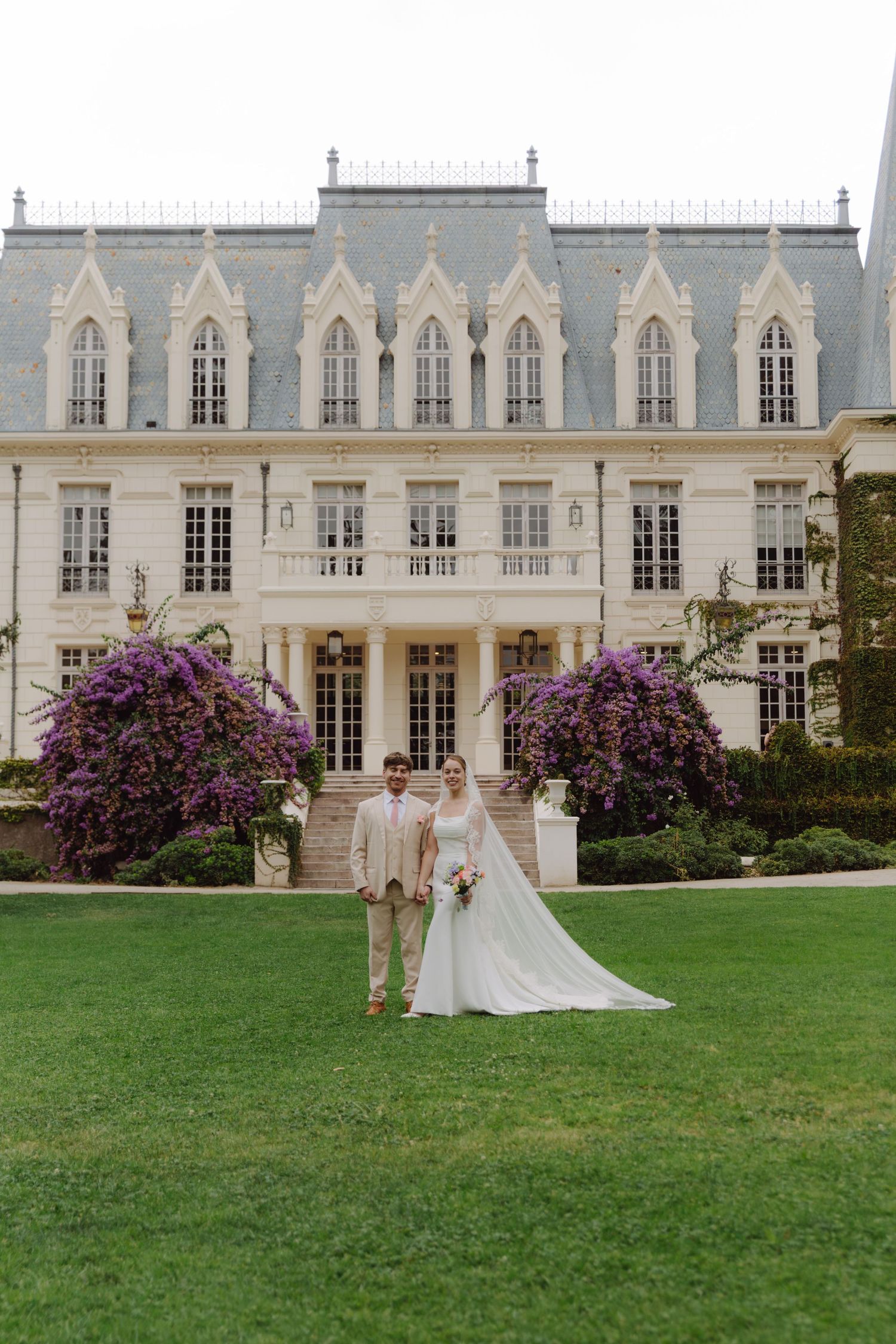 Wedding couple stands on green lawn in front of an elegant French chateau with purple blooms.