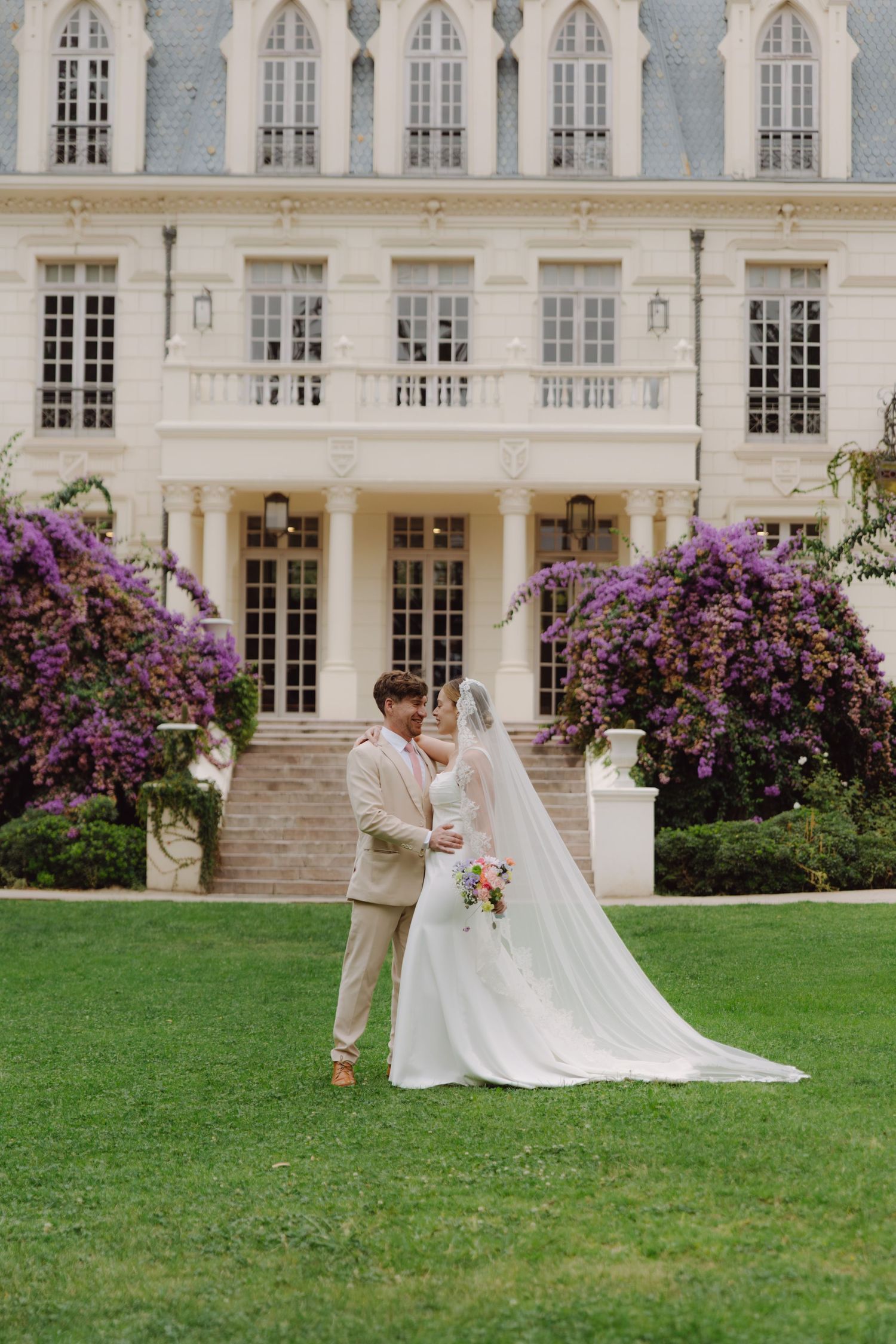 Newlyweds embrace on a manicured lawn before a grand chateau adorned with purple bougainvillea.