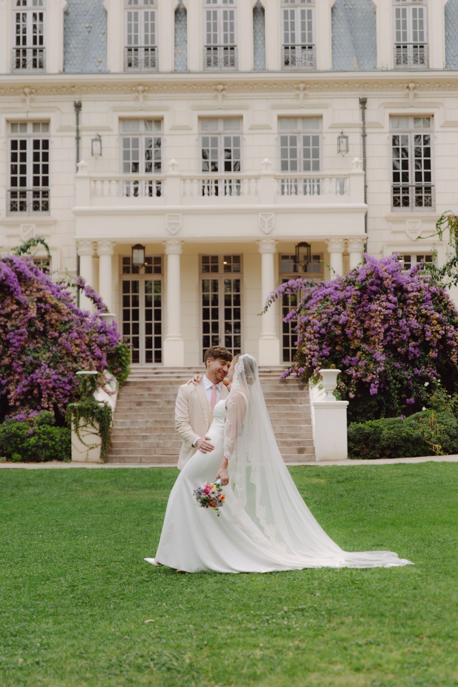 Bride and groom stand close together on lush grass before a stately mansion with purple flowers.