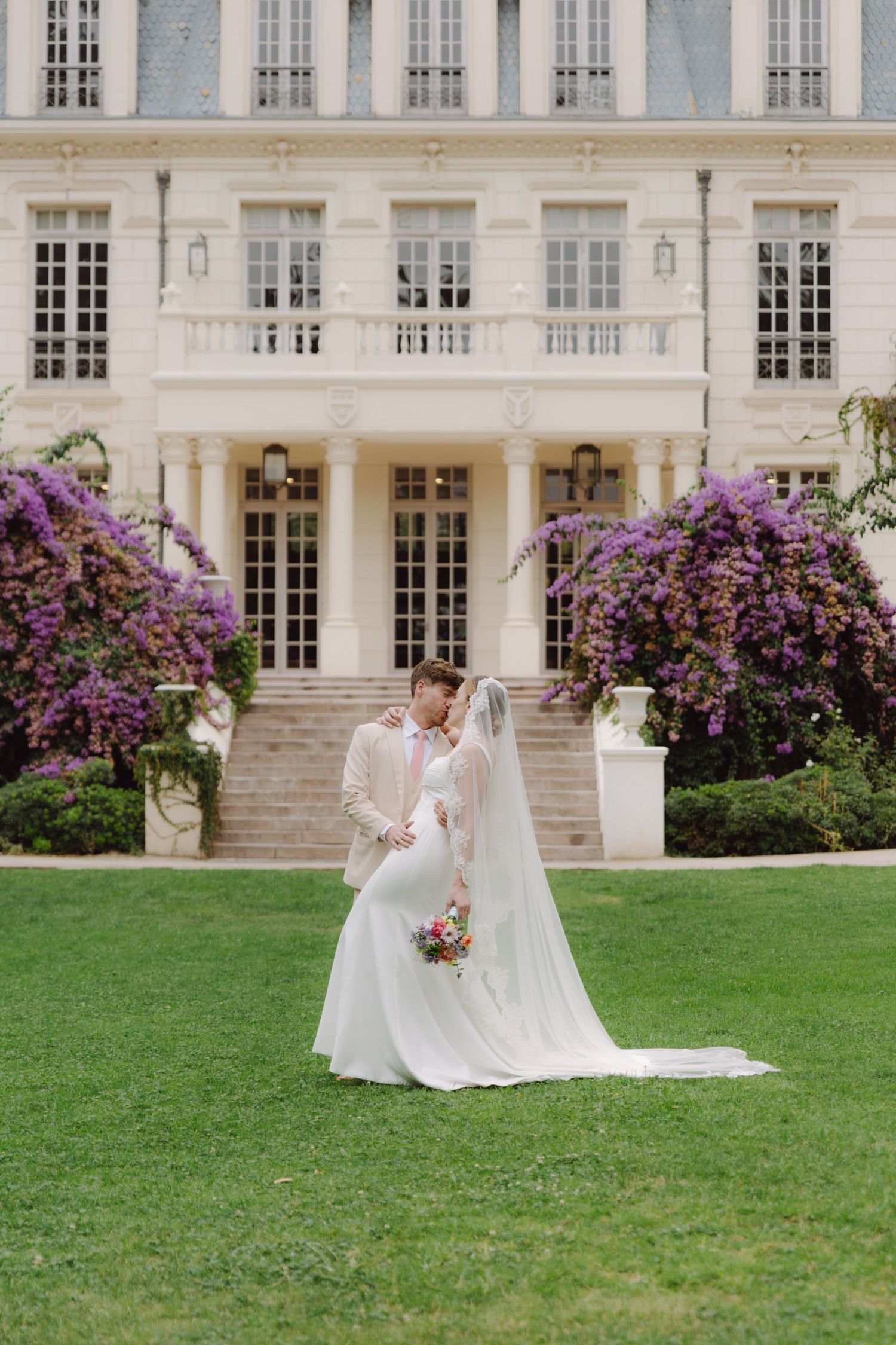 Romantic wedding couple leans in for a kiss on a green lawn before a French chateau.