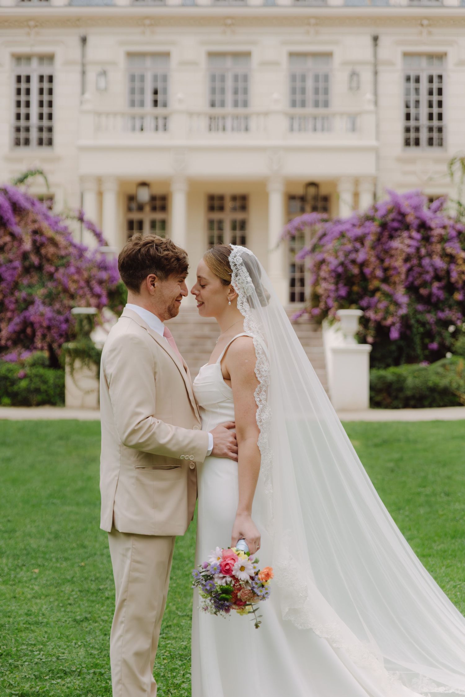 Bride and groom share an intimate moment on a garden lawn before a grand estate with purple blooms.