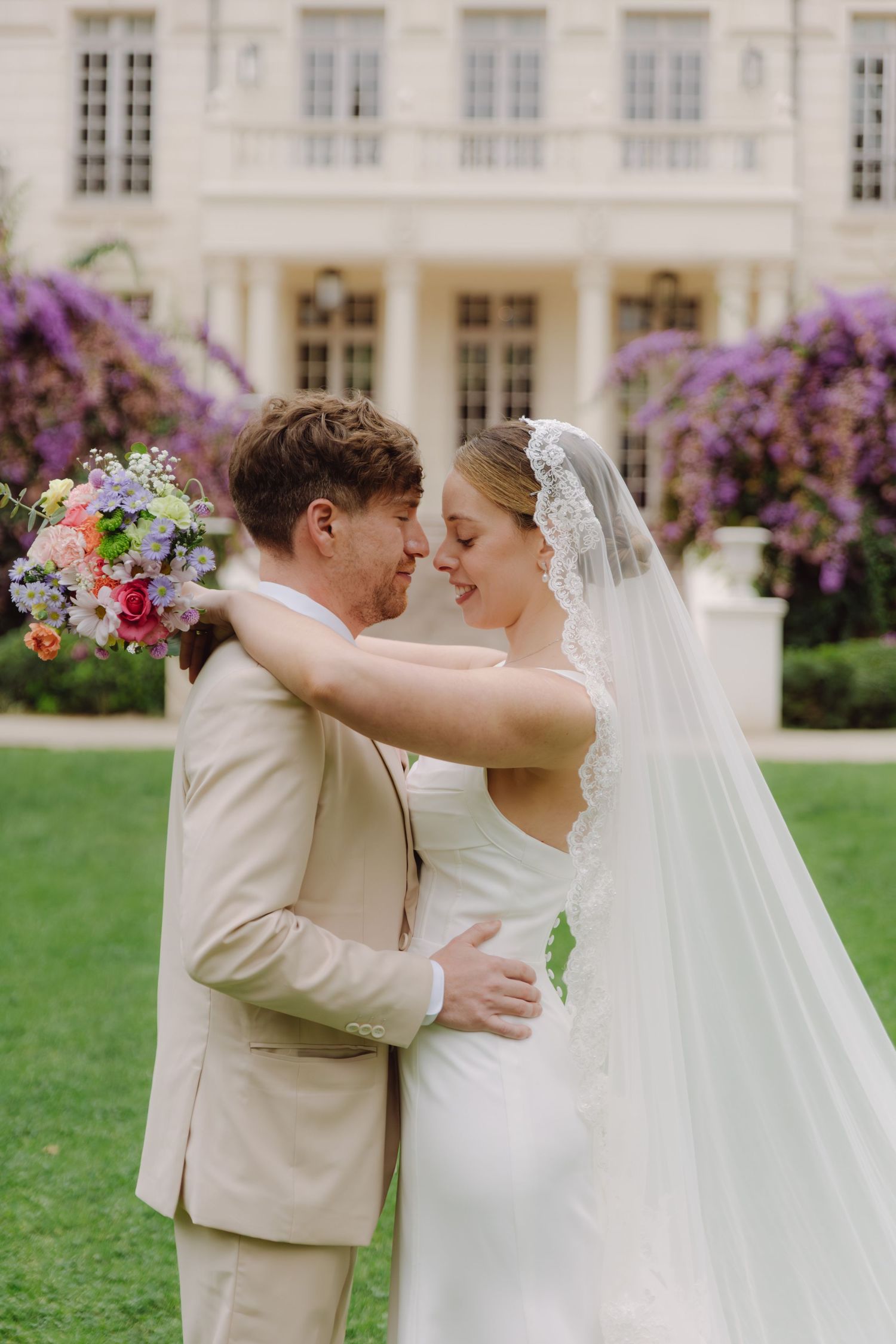 Newlyweds embrace and smile at each other holding a colorful bouquet before a grand French manor.