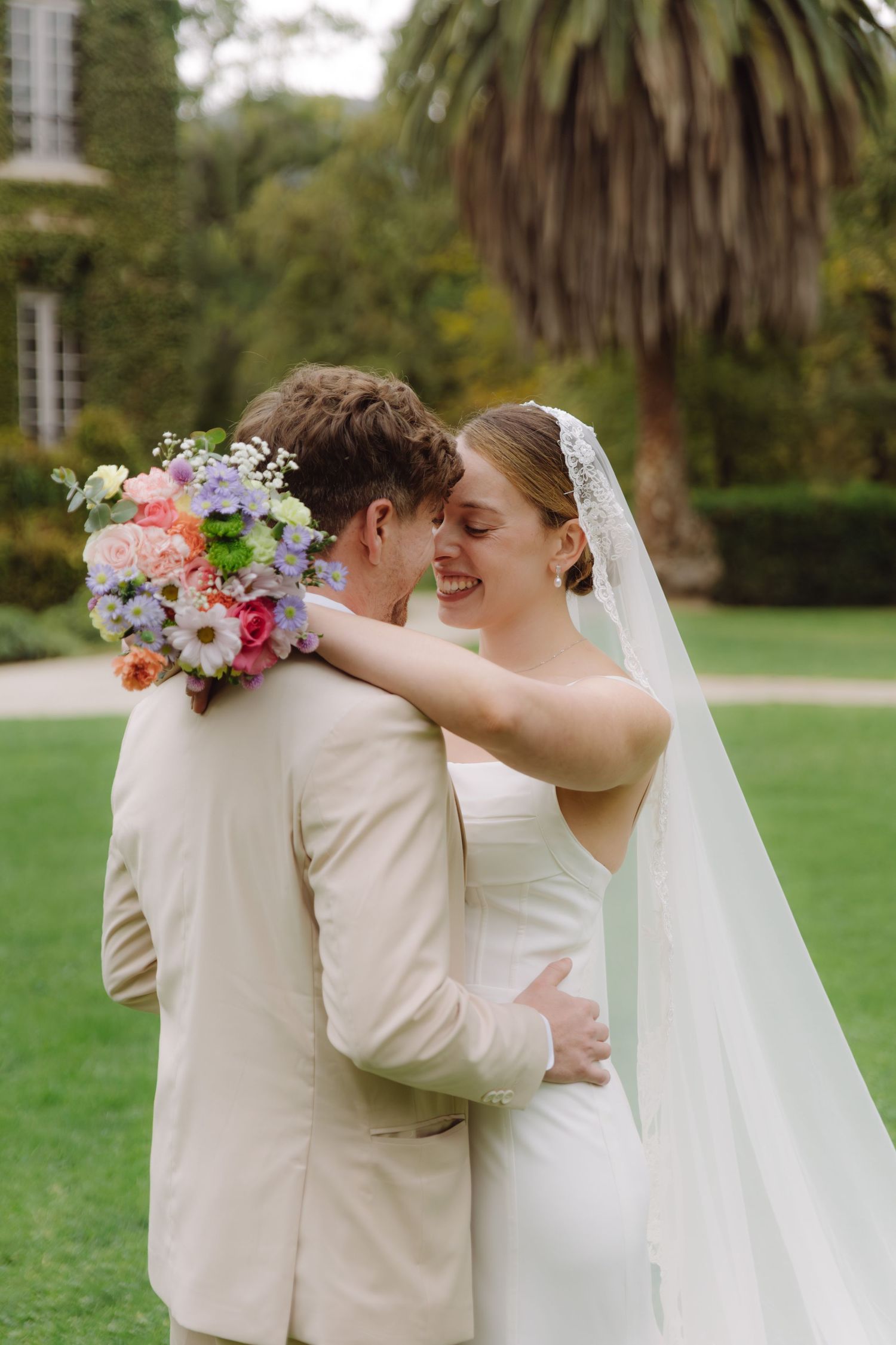 Bride and groom embrace on green lawn, bride smiling with colorful bouquet and lace veil at outdoor wedding.