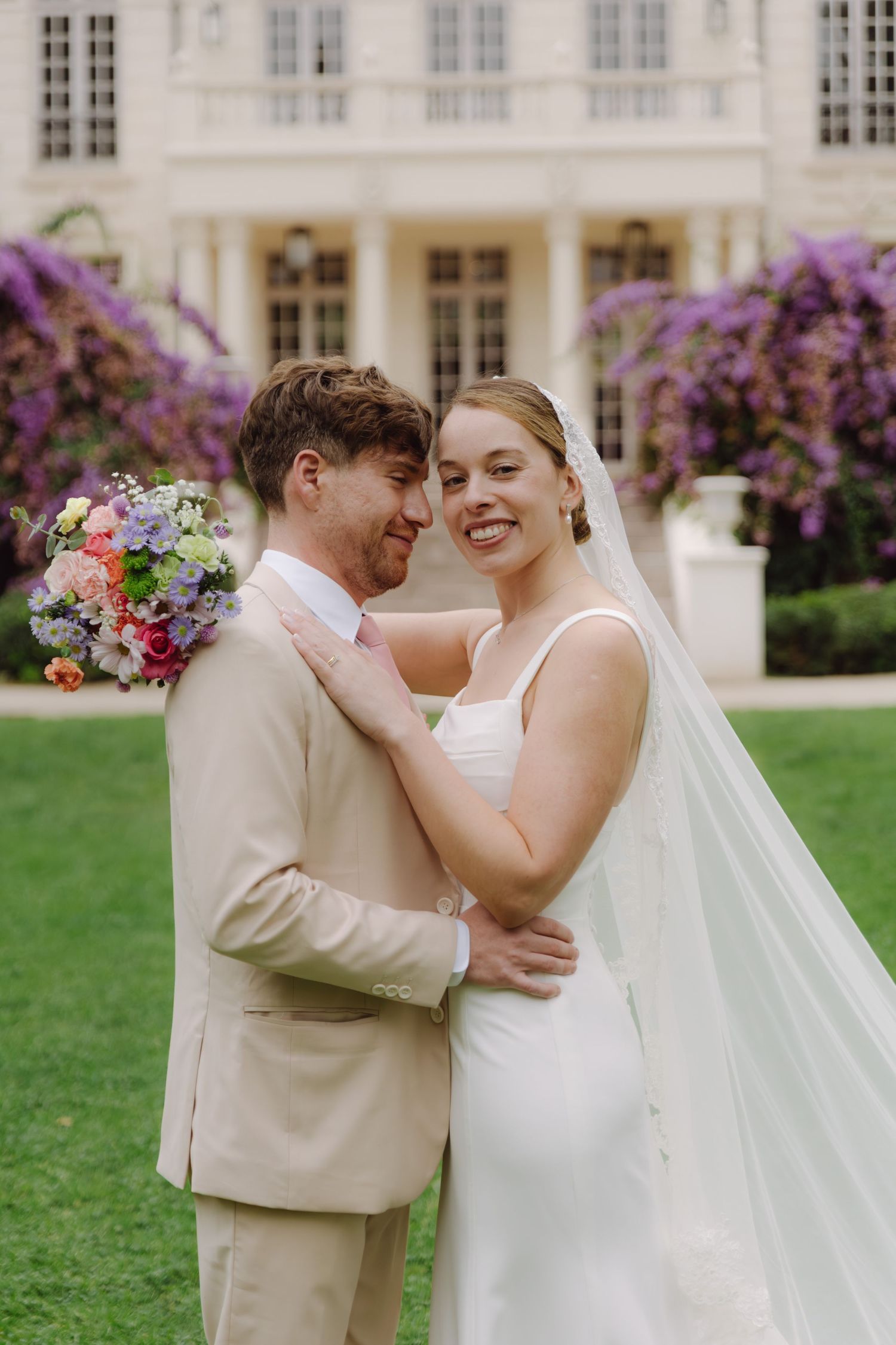 Smiling bride in white gown and veil embraces groom in beige suit before elegant mansion with purple flowers.