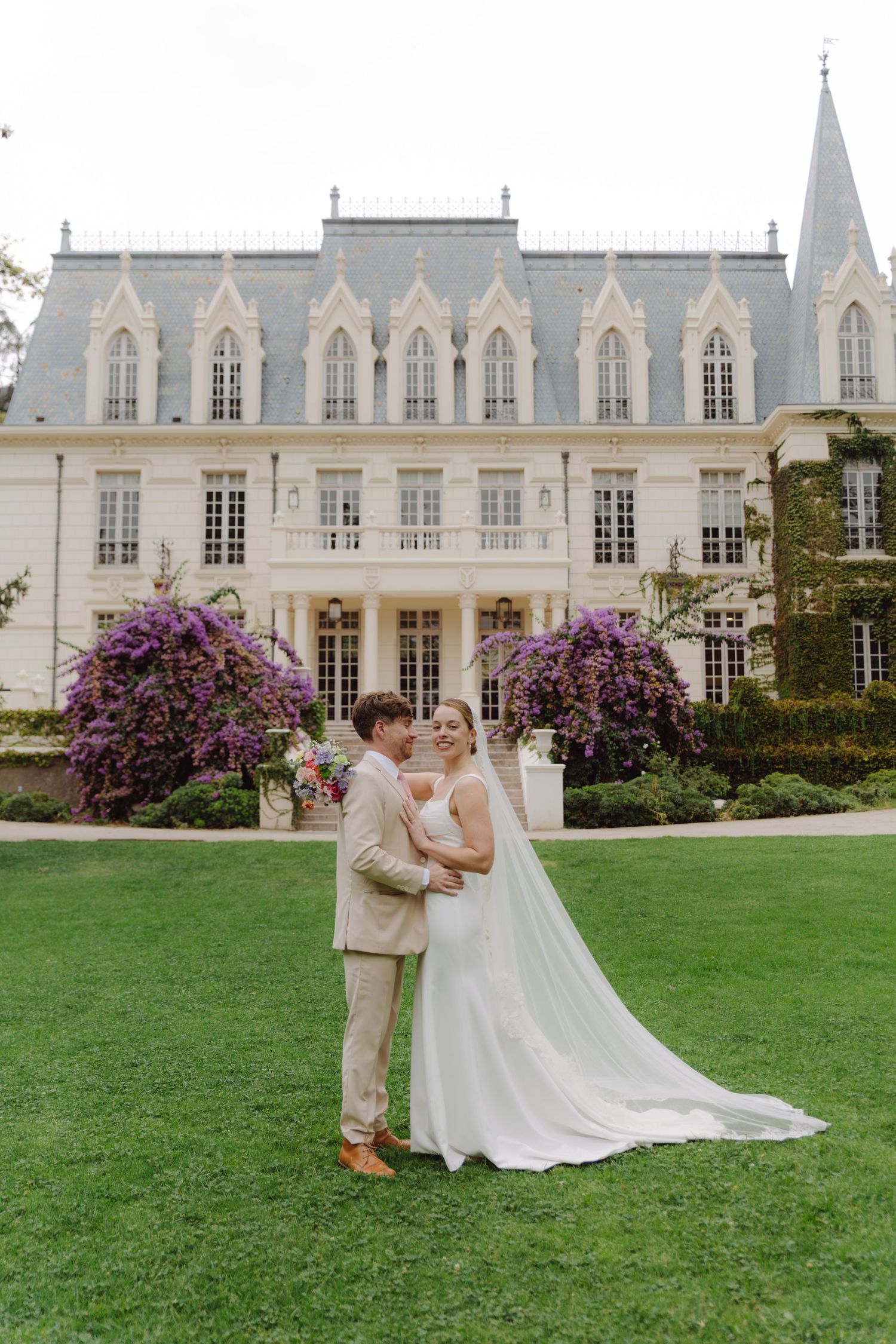 Bride and groom pose on lawn before grand Gothic-style chateau with purple flowering trees on wedding day.