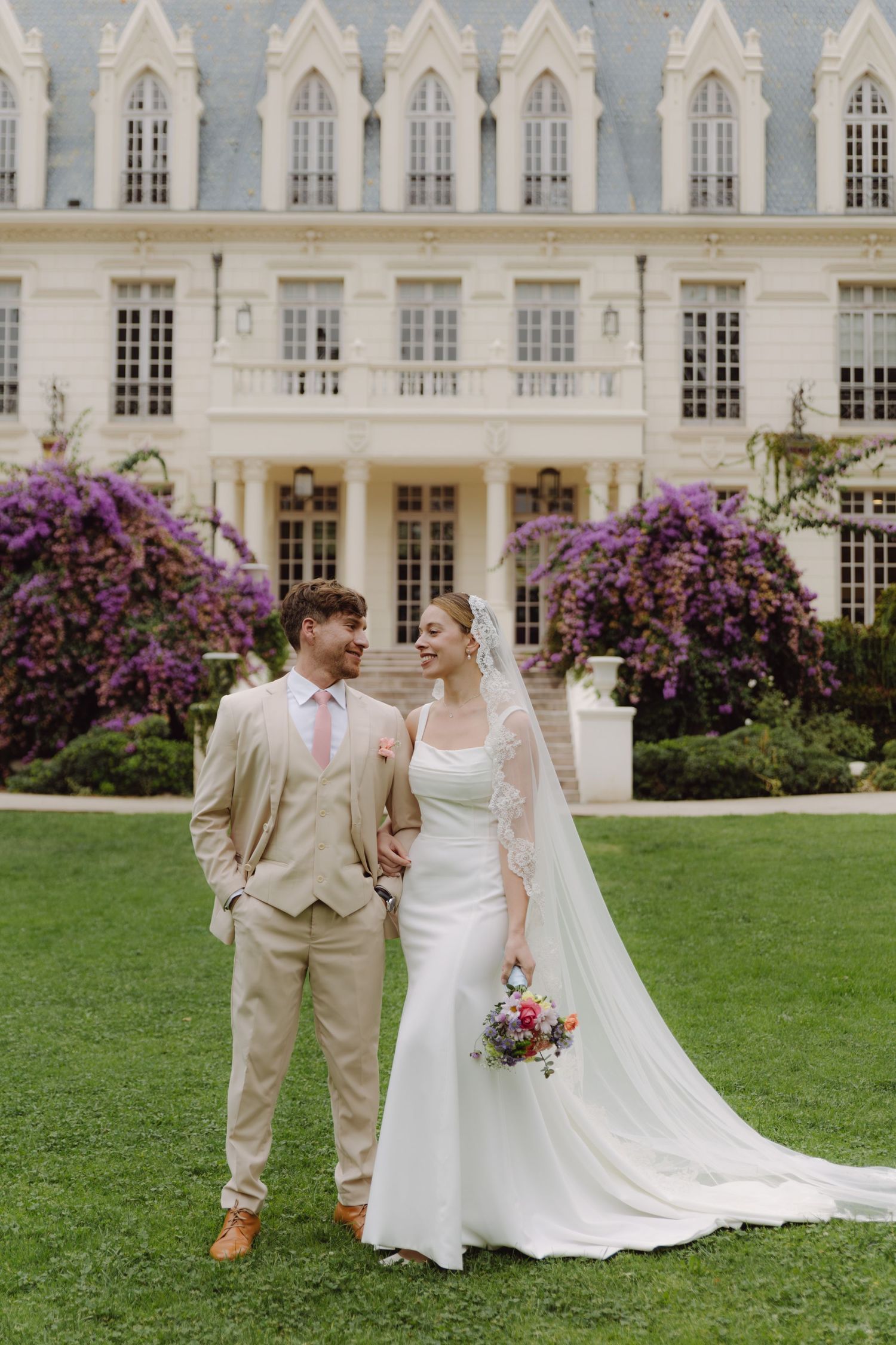 Bride holding colorful bouquet and groom in beige suit walk hand in hand before ornate chateau with purple blooms.