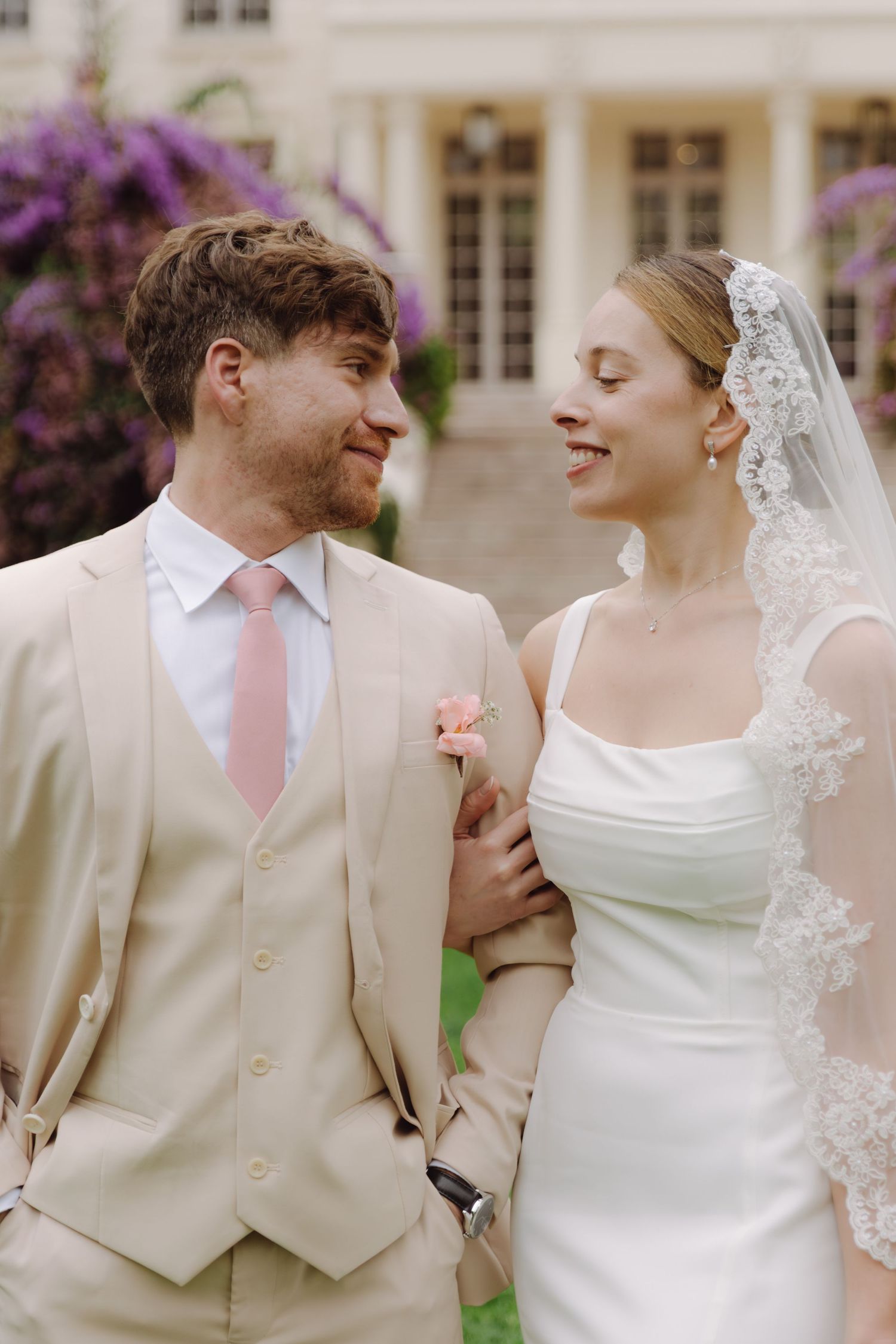 Bride in lace veil and groom in beige suit with pink tie share a loving glance at wedding venue.