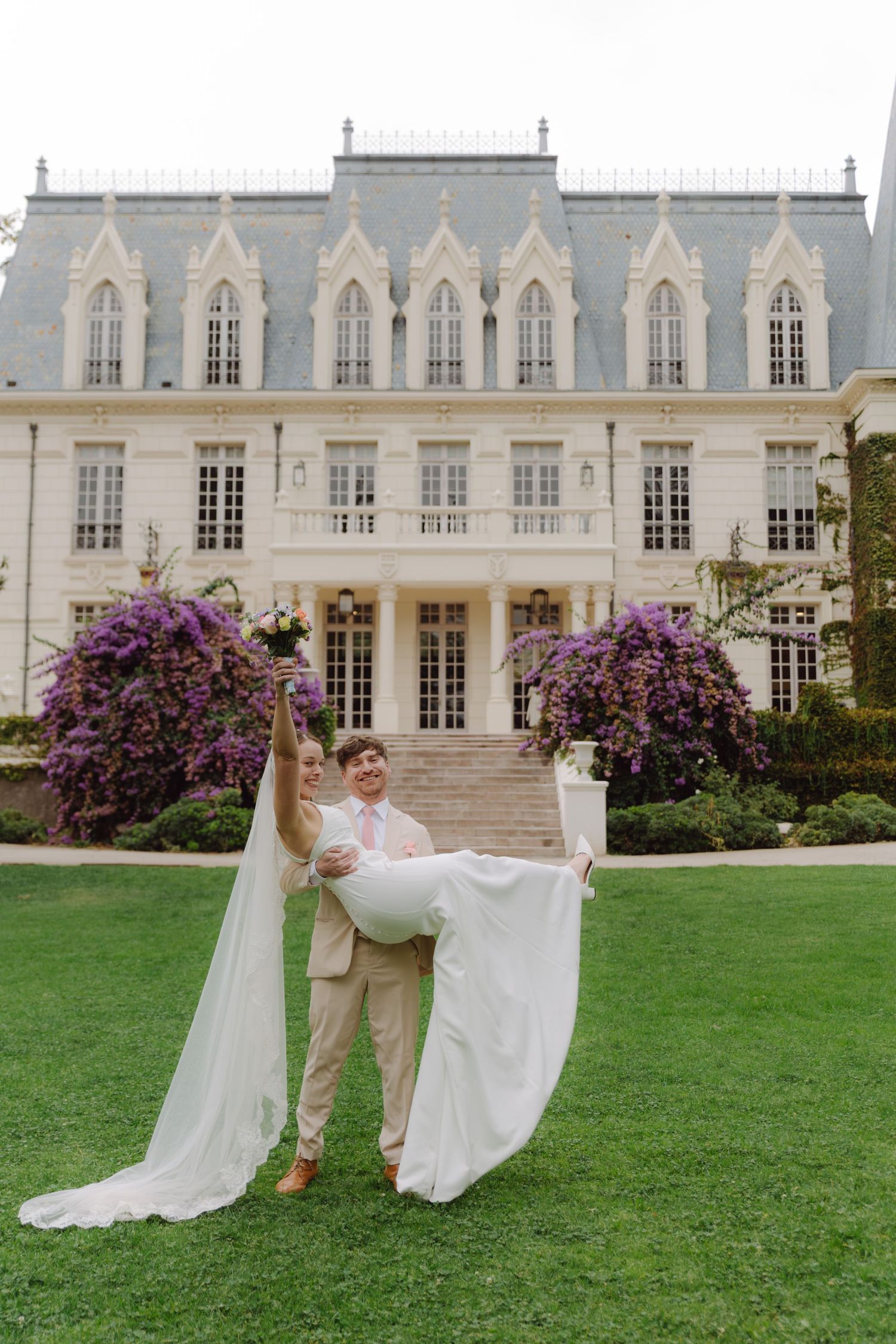 Groom in beige suit lifts laughing bride in white gown before grand Gothic mansion with purple flowering trees.