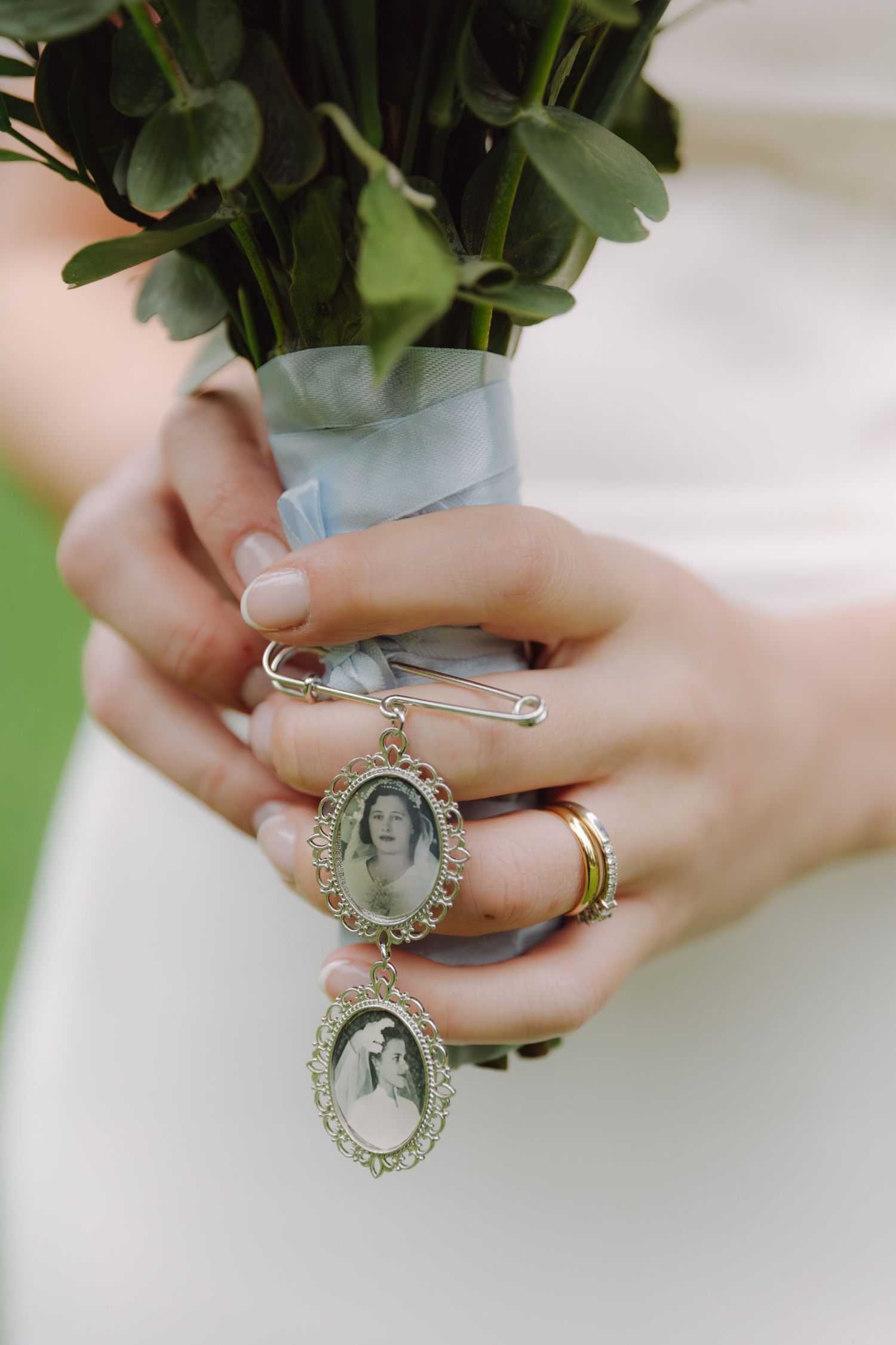 Close-up of bride holding bouquet with vintage oval photo locket charms pinned as sentimental wedding keepsake.